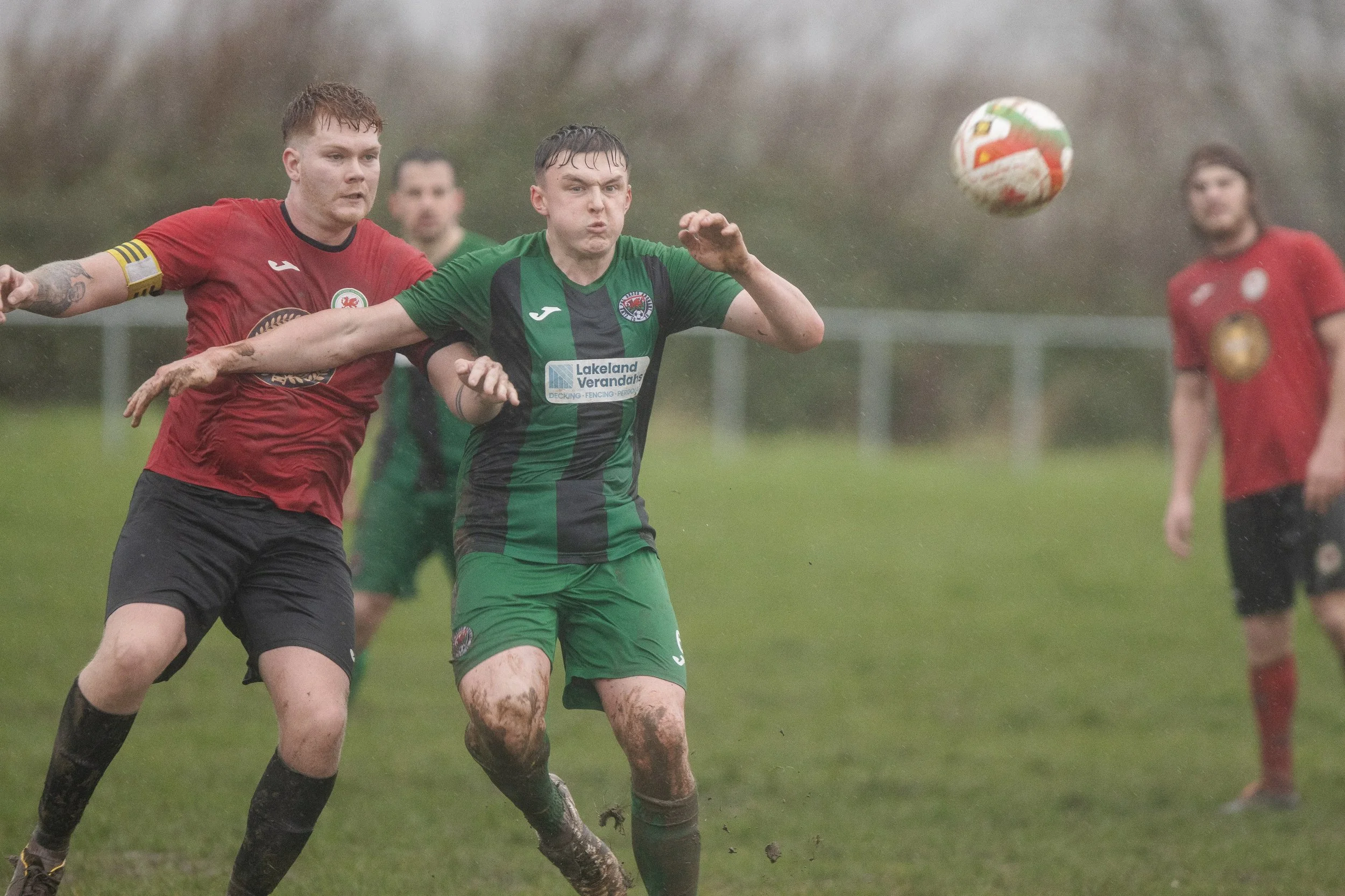Two soccer players, one in a red jersey and the other in a green jersey, compete for the ball on a rainy, muddy field, with two other players in the background.