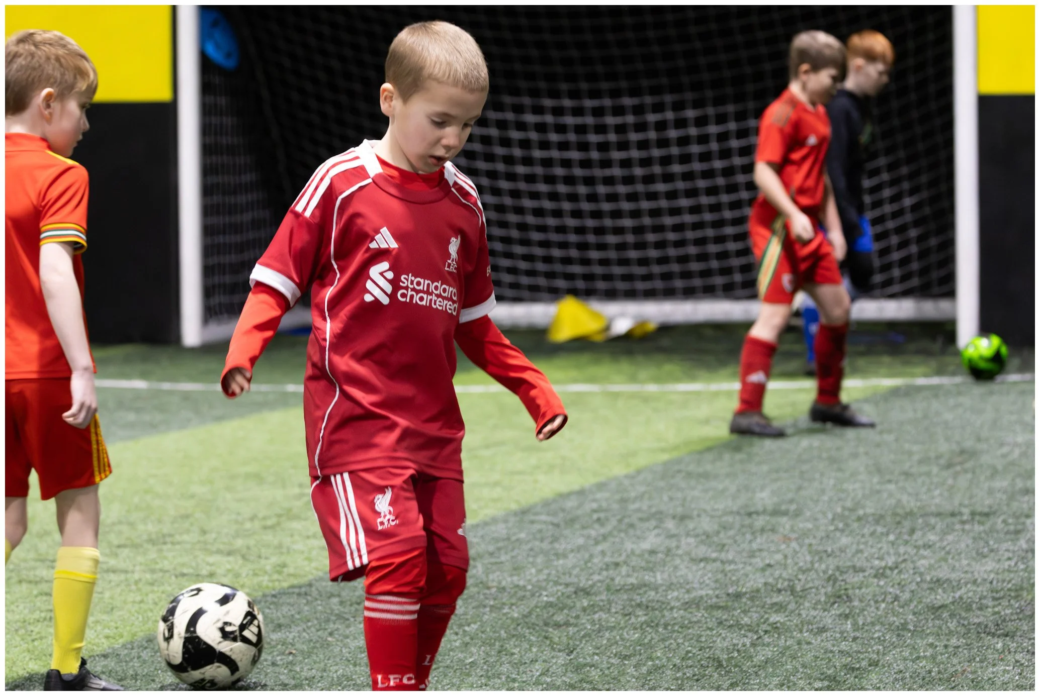 Young boys playing indoor soccer on a field, one in a Liverpool jersey, standing with a soccer ball, others near the goalpost, on artificial turf.