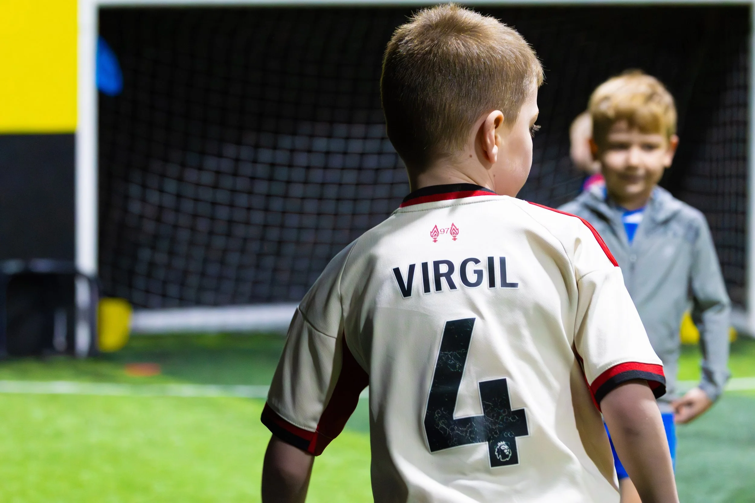 Two young boys on an indoor soccer field, one wearing a white jersey with the name 'Virgil' and the number '4' on the back, facing away, and the other facing towards him, smiling. A black goal net and yellow structure are in the background.