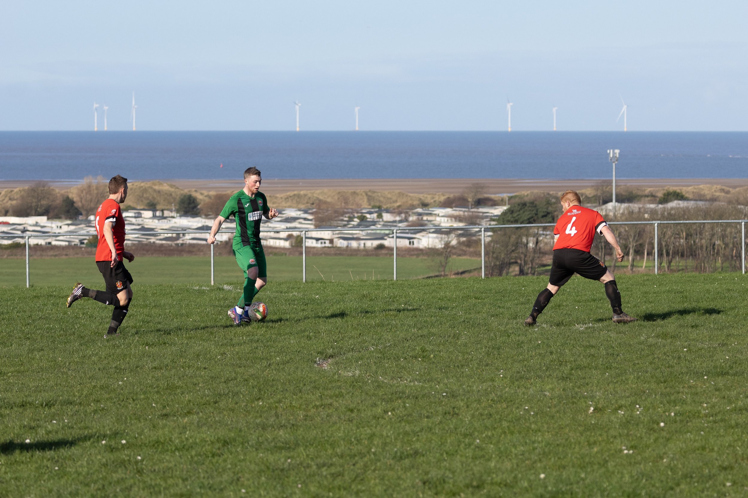 Three soccer players on a grassy field, with a fence, coastal area, and wind turbines in the background.