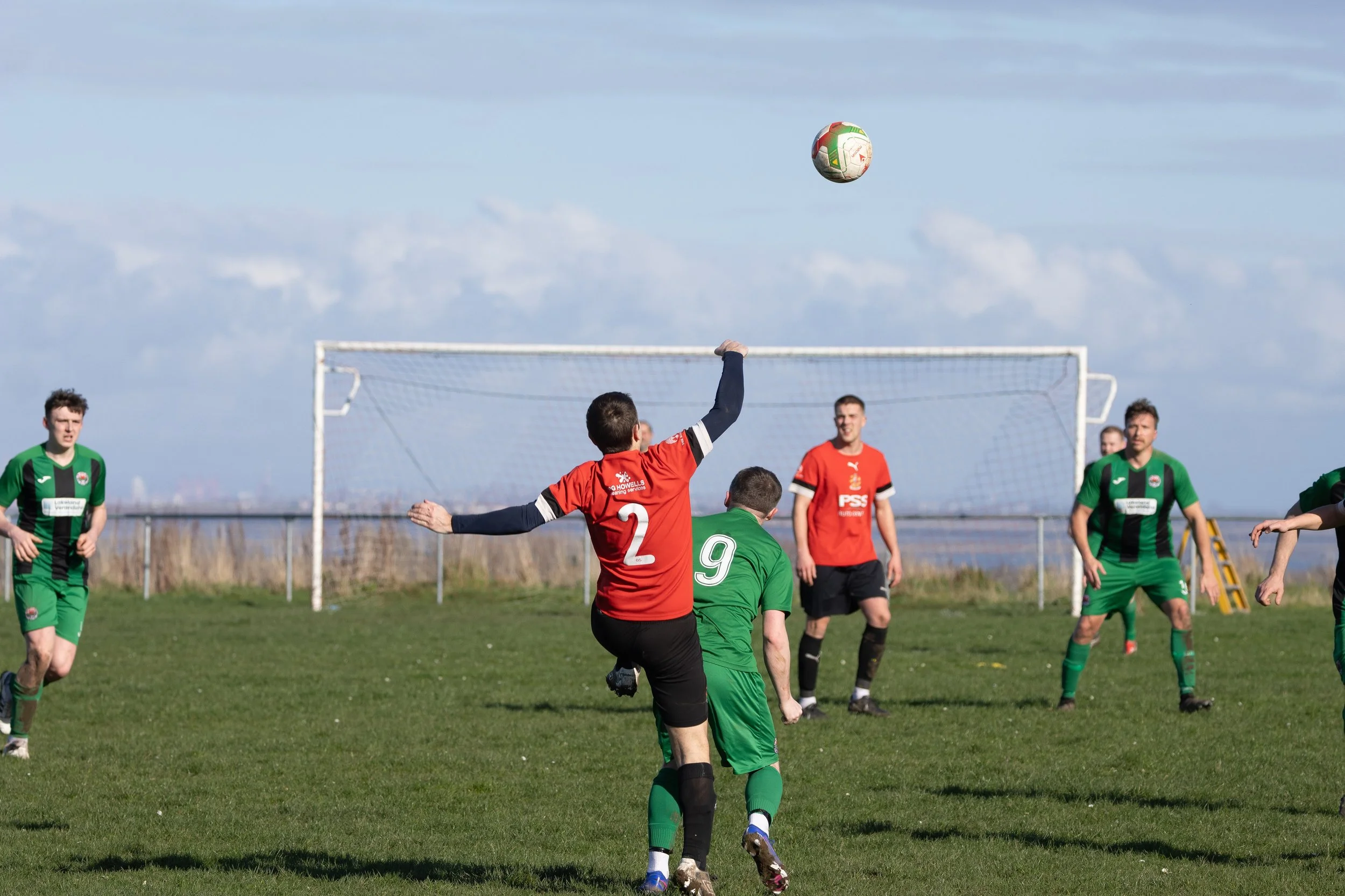 Soccer match with players in red and green jerseys on a field, a player in red jumps to head the ball while others watch.