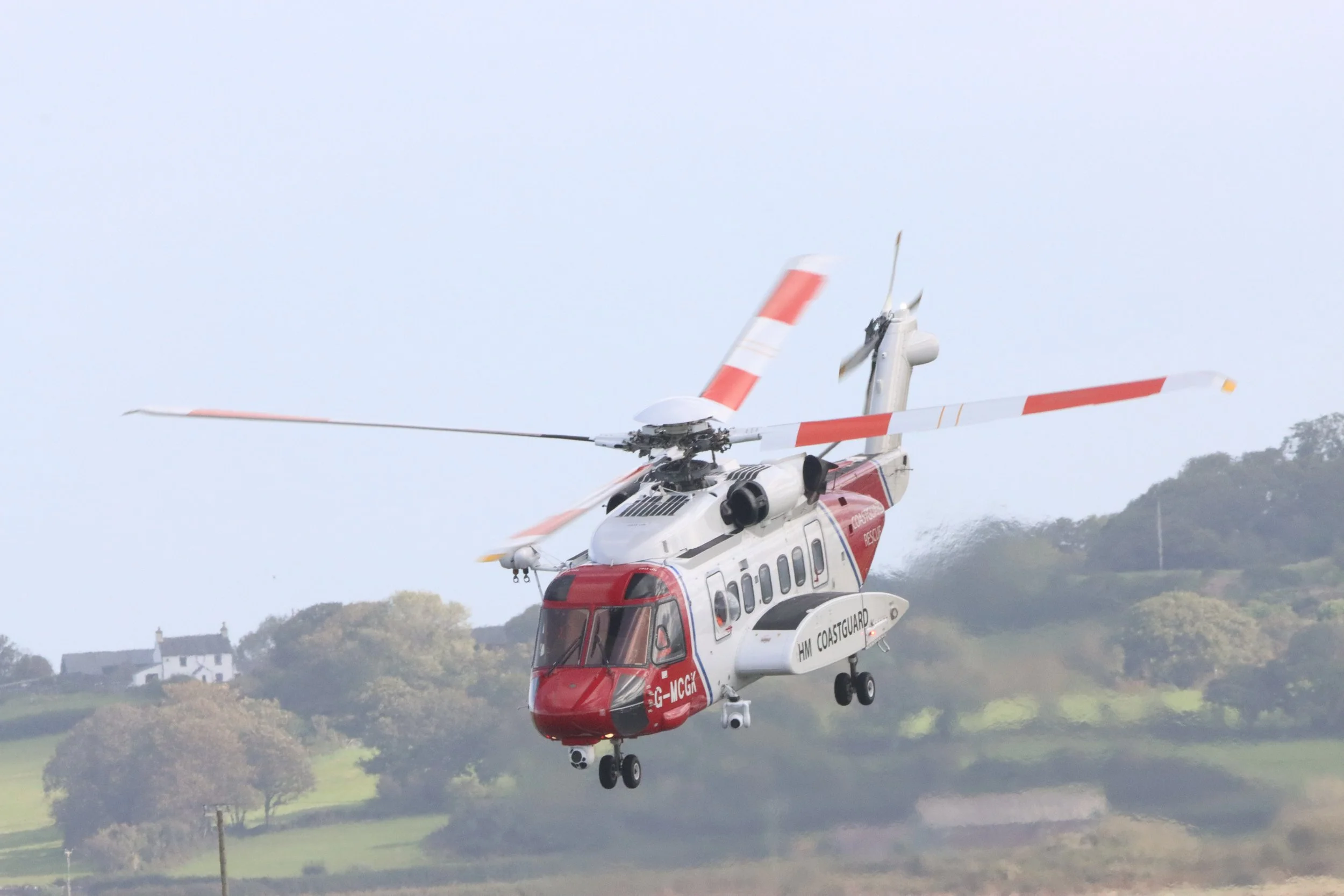 A rescue helicopter flying above a green landscape with trees and a small white building, with hills in the background.