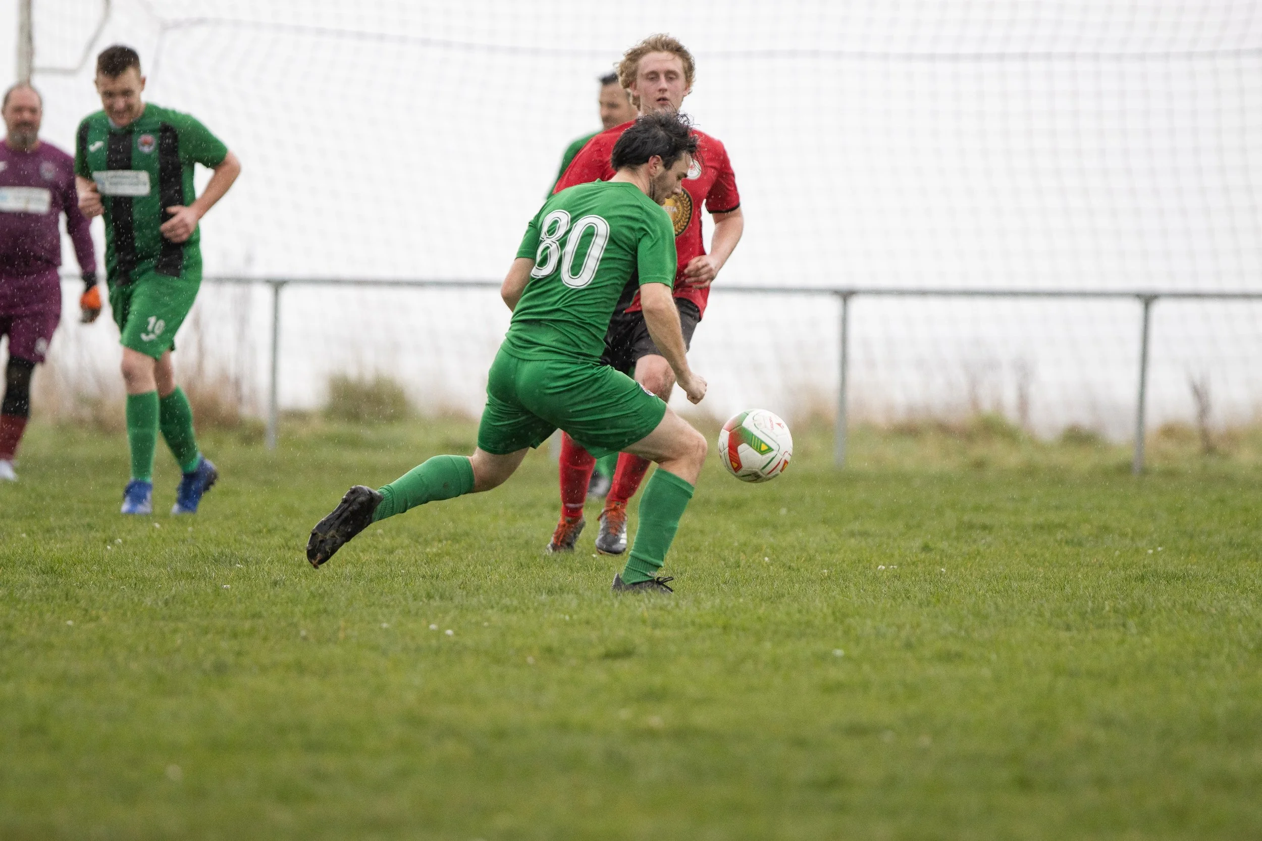 Soccer players in green and red jerseys competing for the ball on a grassy field in the rain.
