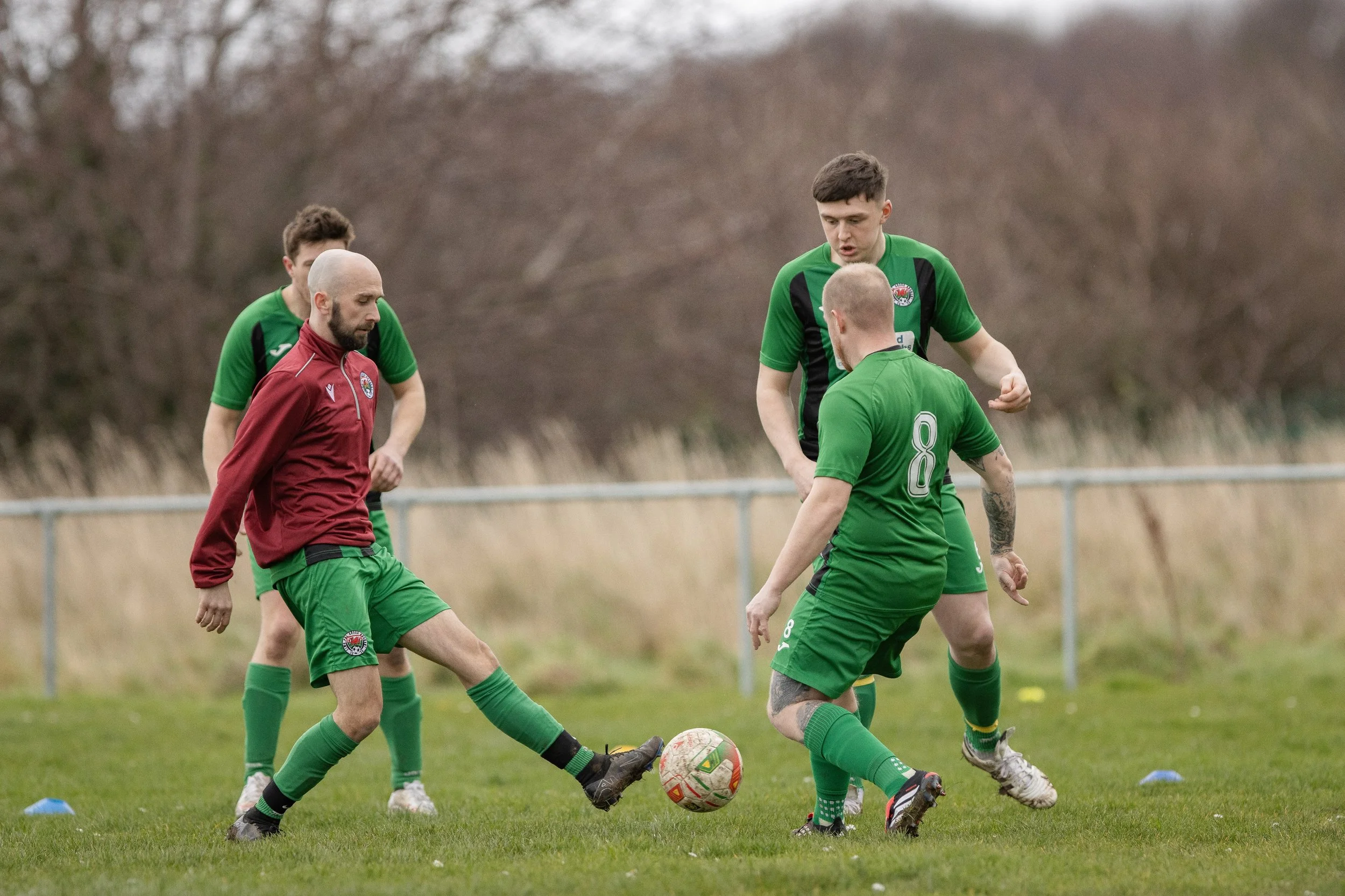 Four men playing soccer on a grassy field, two in green jerseys, one in red jacket, and one mostly obscured. One green player is kicking the ball while the others watch and prepare to respond.