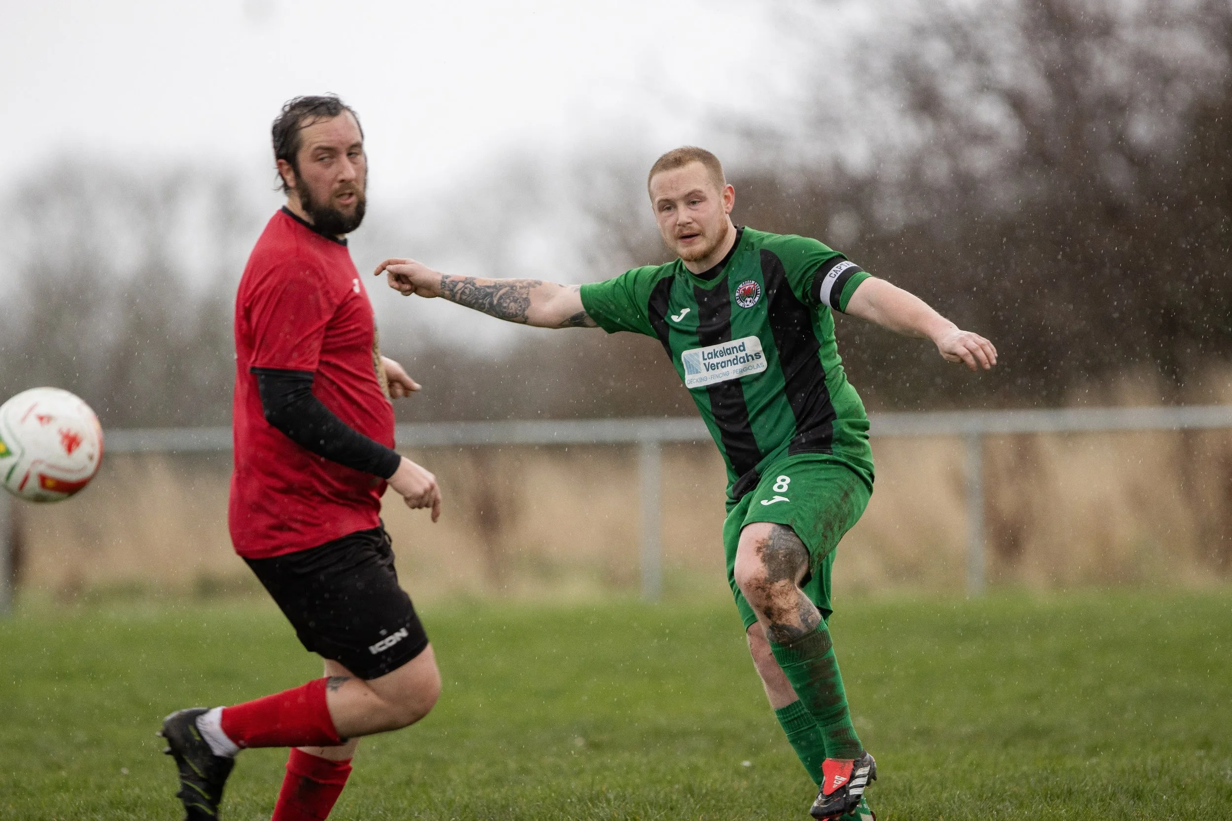 Two soccer players competing for the ball during a match in rainy weather, one in a red uniform and the other in a green and black uniform.