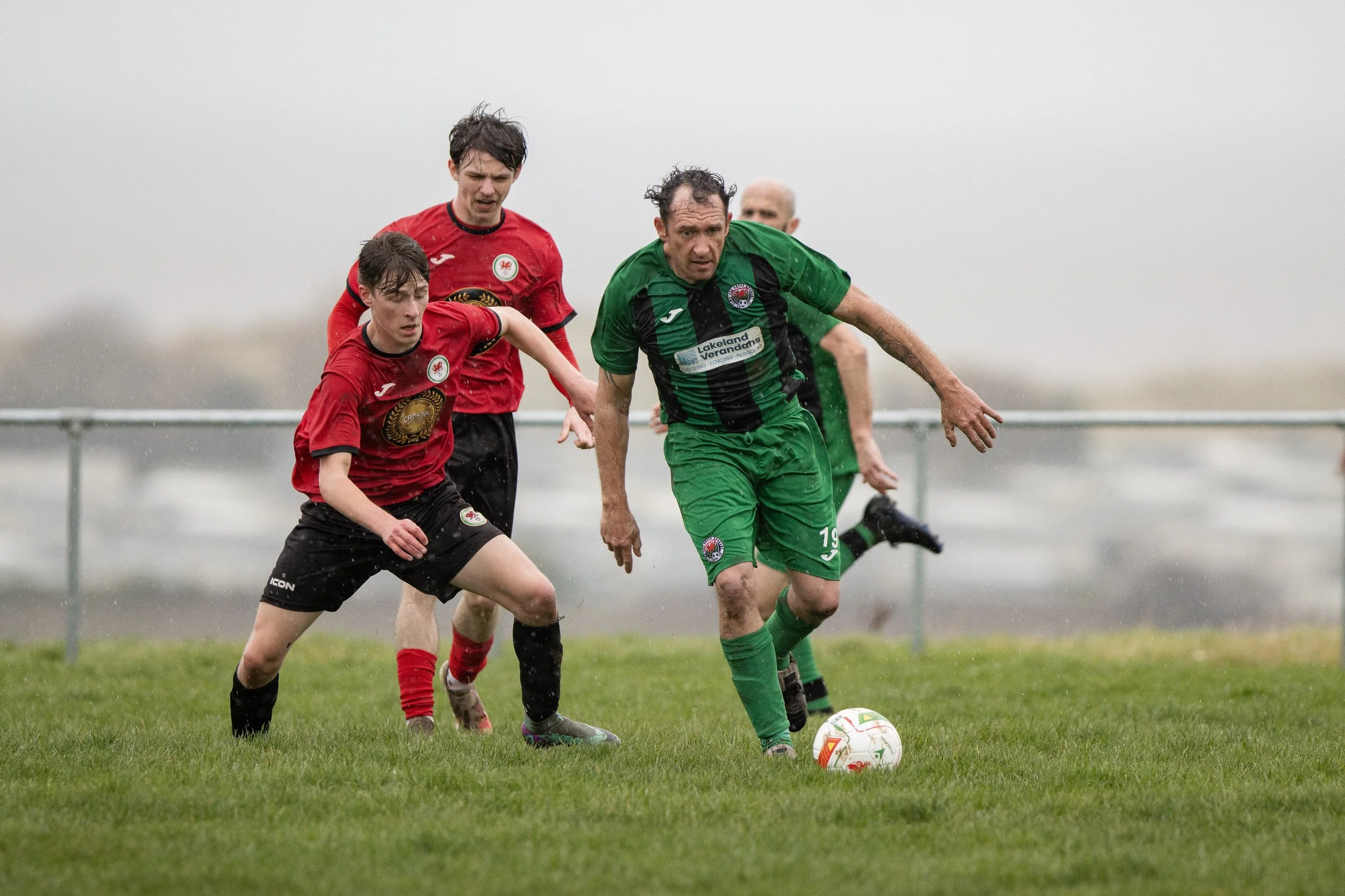 Soccer players competing for the ball on a rainy day, with one player in green and the others in red.