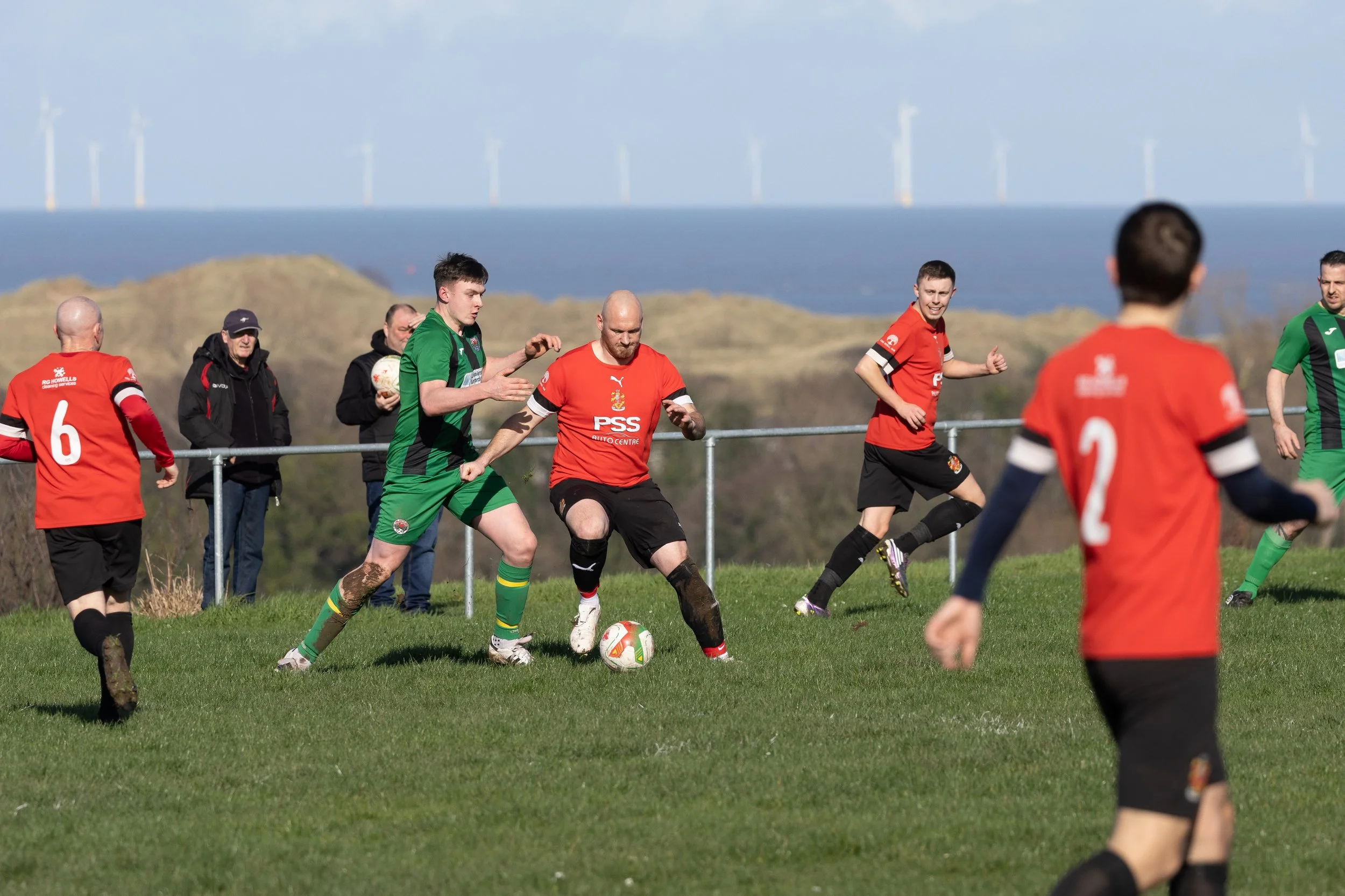 Soccer players in red and green jerseys playing on a grassy field near the coast, with wind turbines on the horizon.