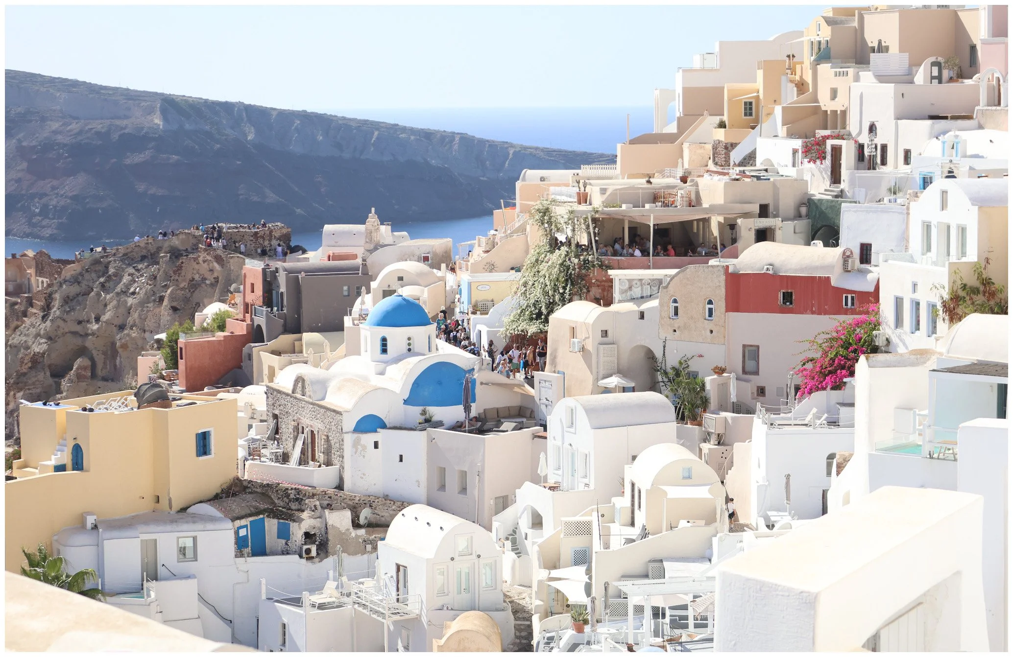 A picturesque view of white buildings with colorful accents, including a blue-domed church, in Santorini, Greece, overlooking the sea and volcanic cliffs.