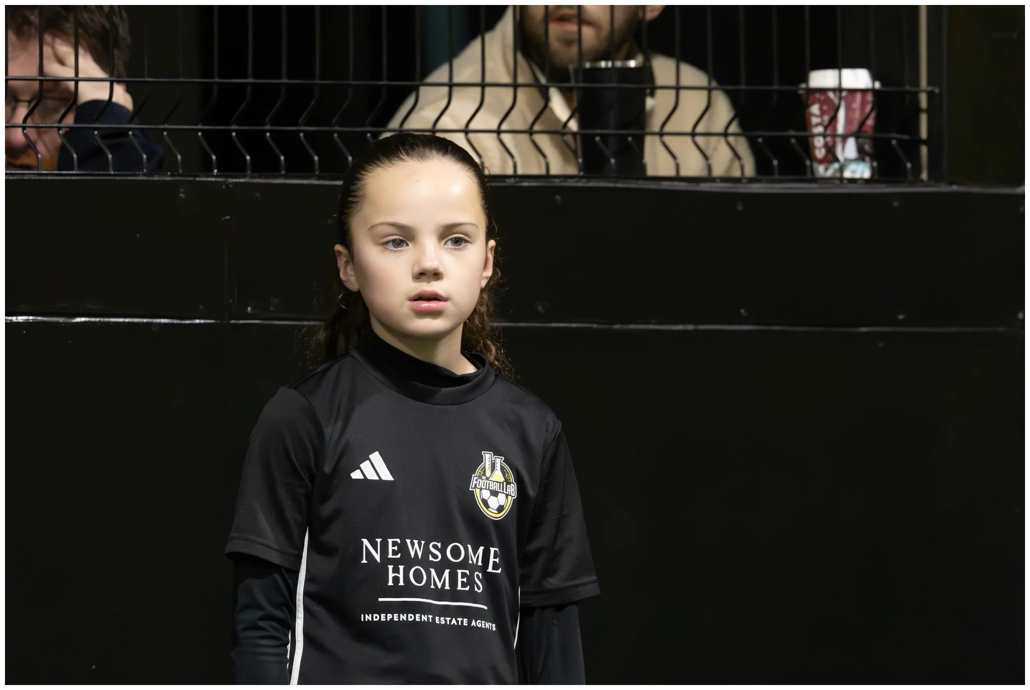 Young girl in black soccer jersey standing on the field with a serious expression, black fence and spectators in the background.