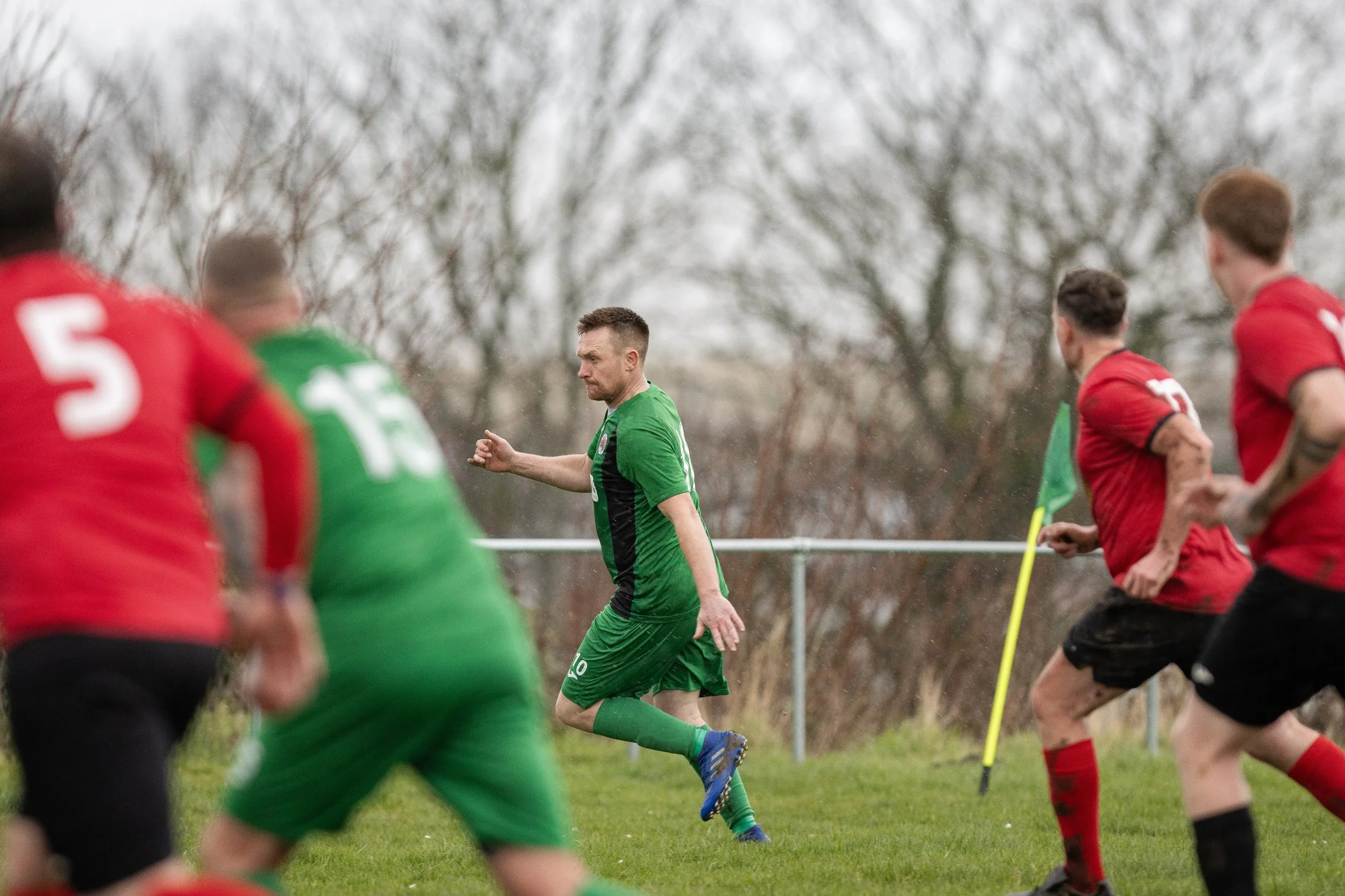 Soccer match with players in red and green jerseys on a grassy field, surrounded by trees in the background, and a player in a green jersey appears to be mid-move.