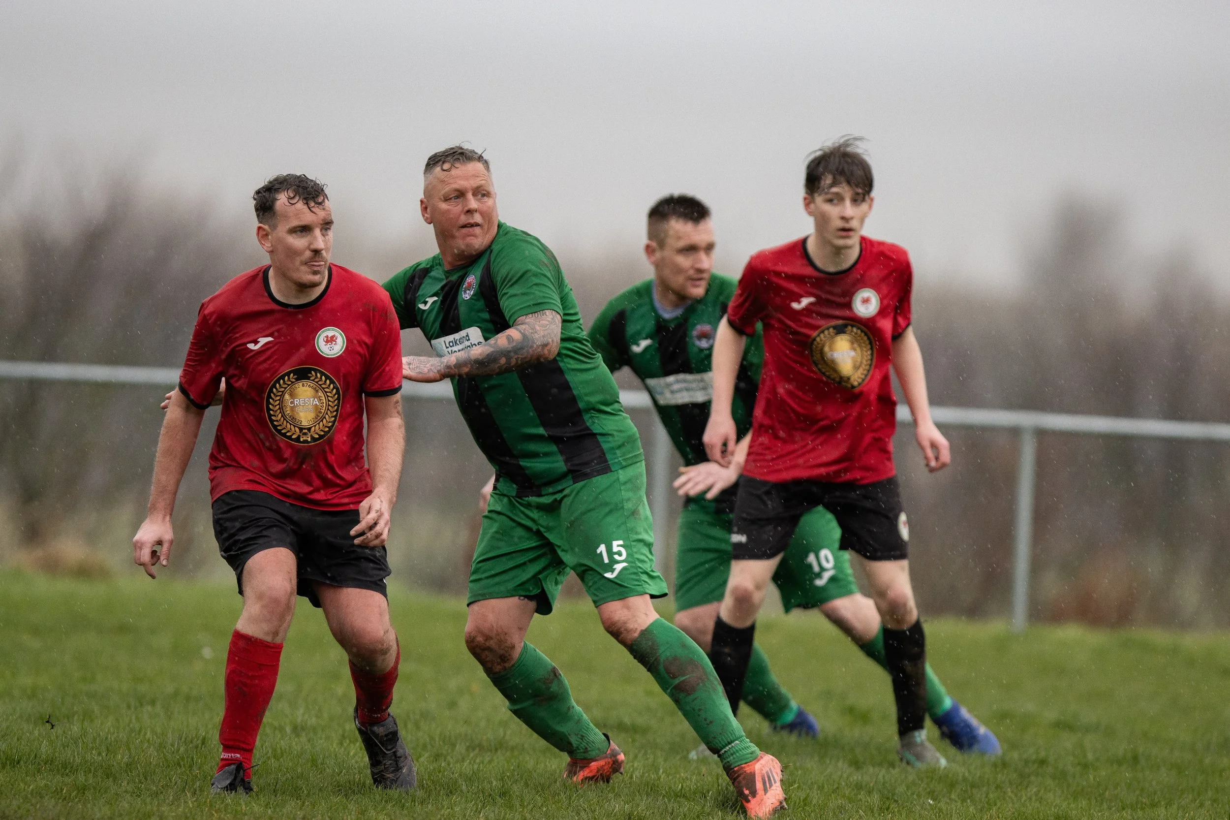 Four male soccer players in red and green uniforms on a rainy field, competing for a ball.