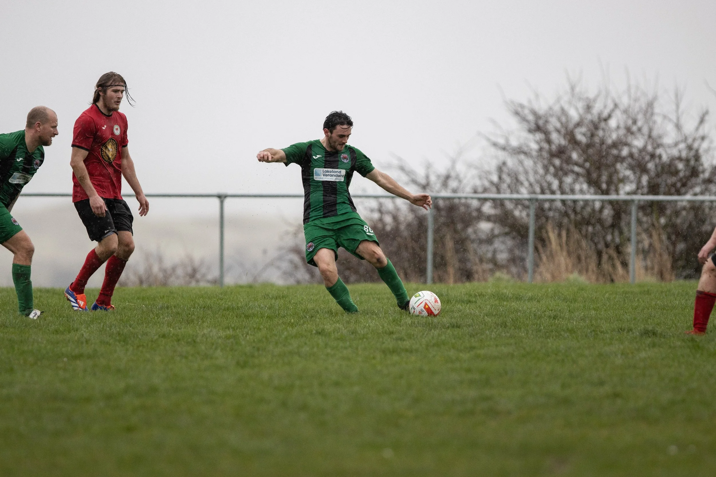 Soccer players on a grassy field during a match, with one player in a green uniform kicking the ball while others in red and black uniforms watch.