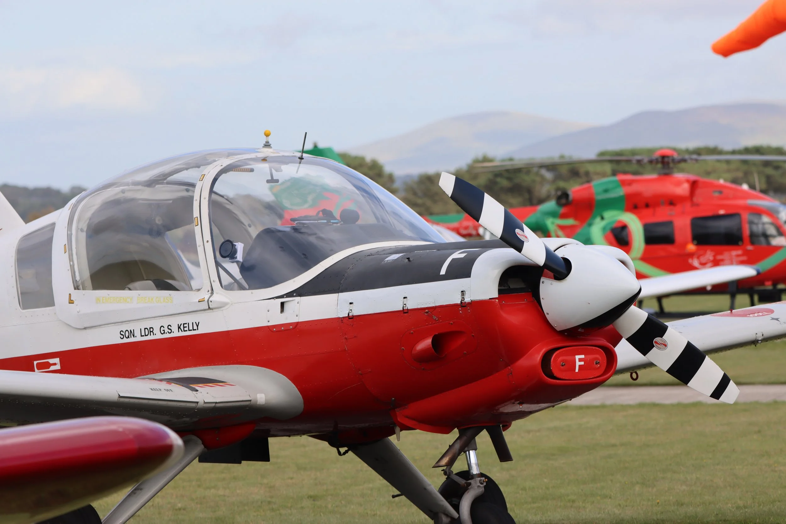 Close-up of a vintage military aircraft with a white, red, and black color scheme, featuring a propeller at the front and a clear canopy over the cockpit, parked on grass with another colorful helicopter in the background and mountains in the distanc