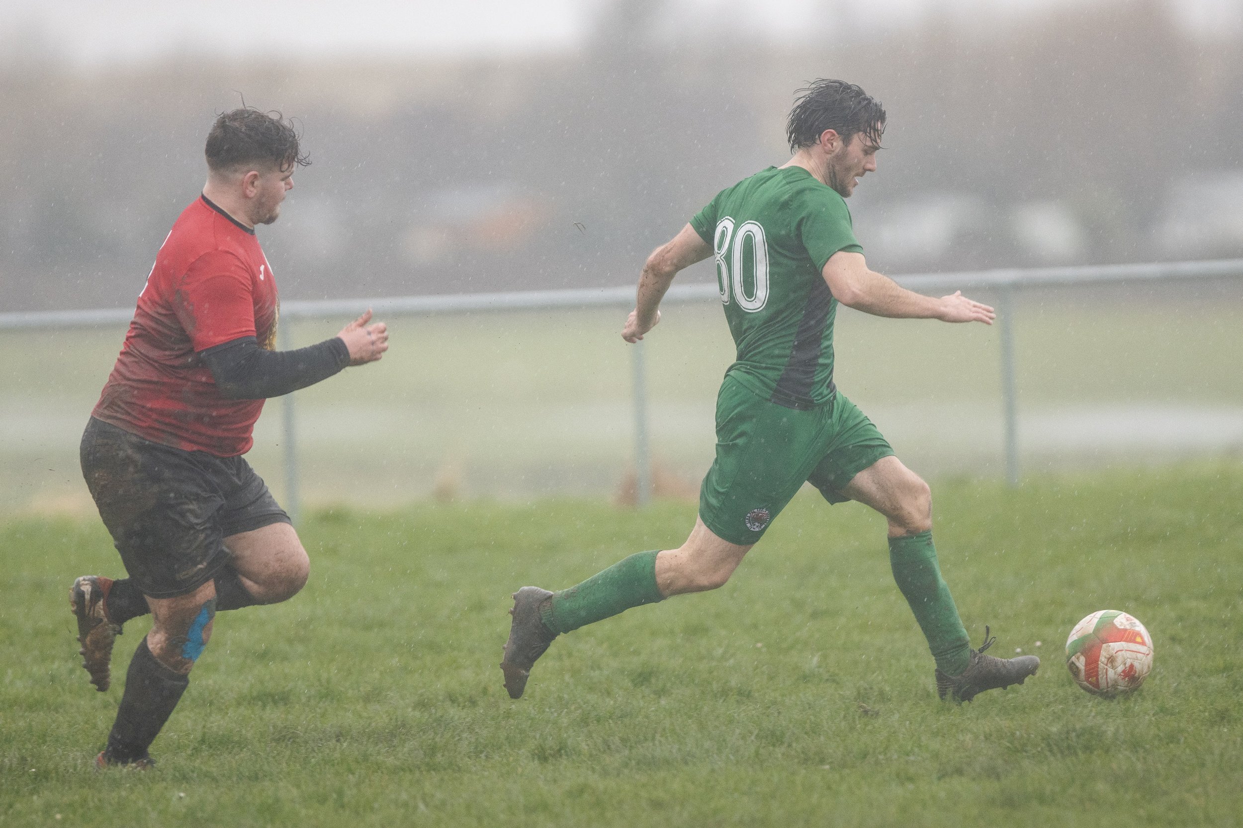 Two soccer players running in the rain on a grassy field, one in a green uniform with number 80 and the other in a red and black uniform.
