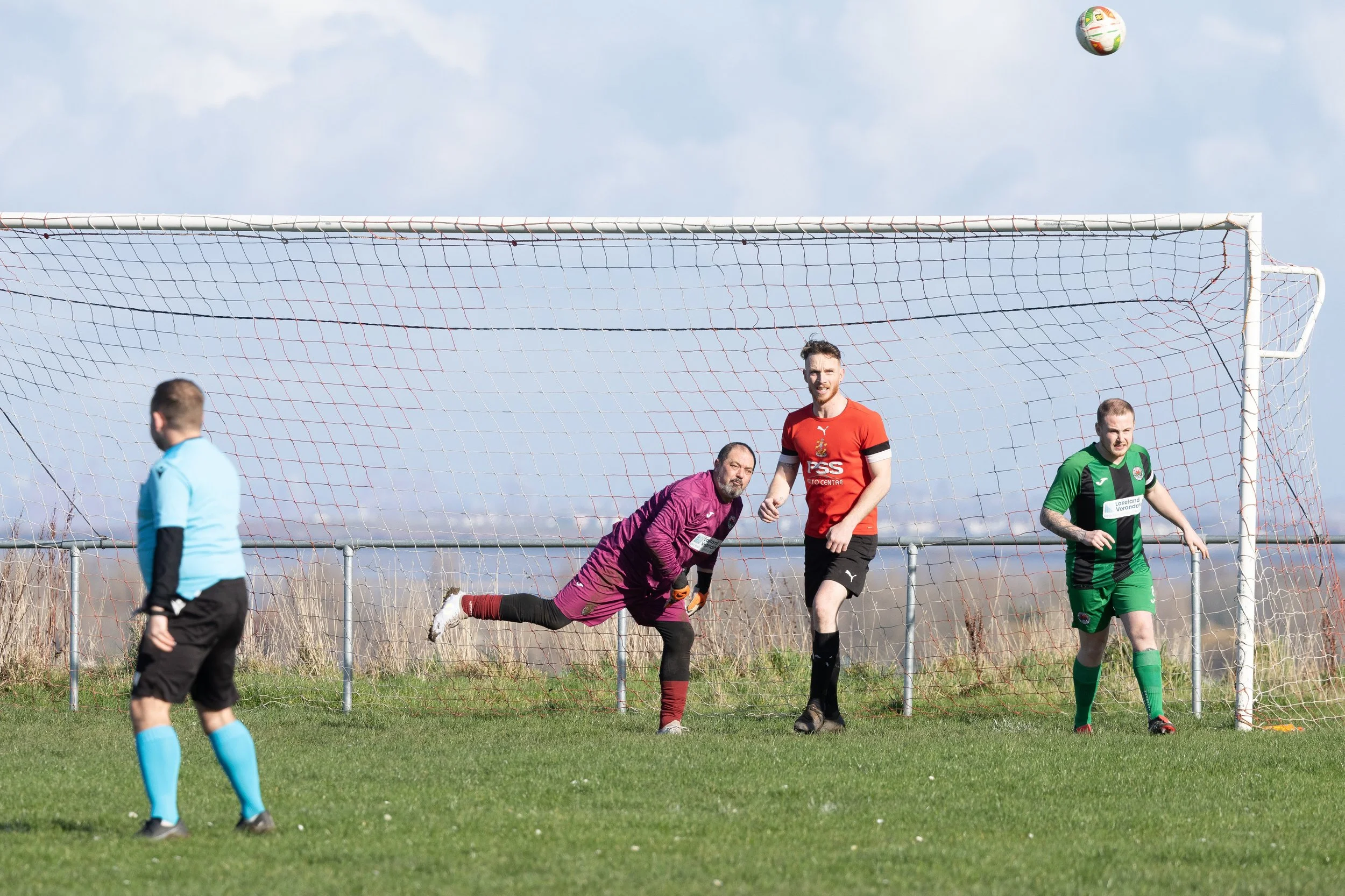 Soccer game with players and referee on the field, one player in a red jersey is close to the goal, a goalkeeper in purple is diving, and another player in green is near the goalpost, with a ball in the air.