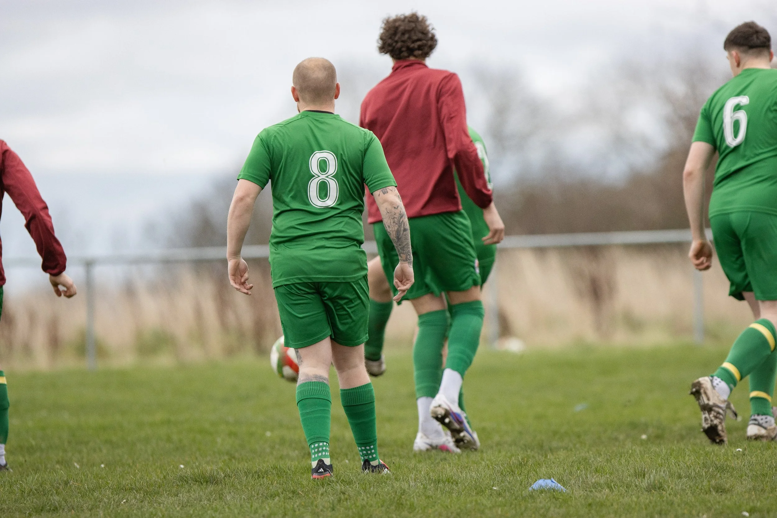 Soccer players in green and red uniforms during practice or game on a grassy field, with equipment and a fence in the background.