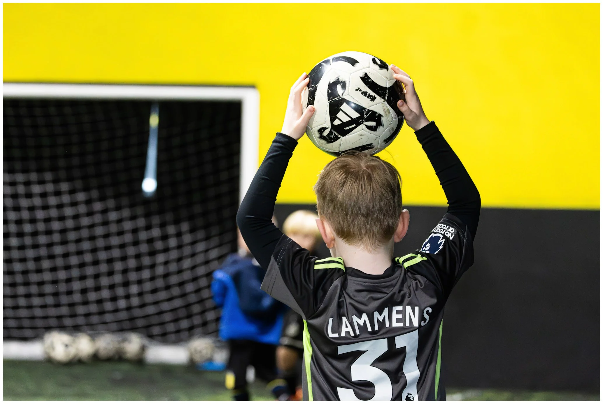 A young boy with short hair in a black sports jersey numbered 31, holding a soccer ball above his head, indoors with a yellow wall and soccer goal in the background.