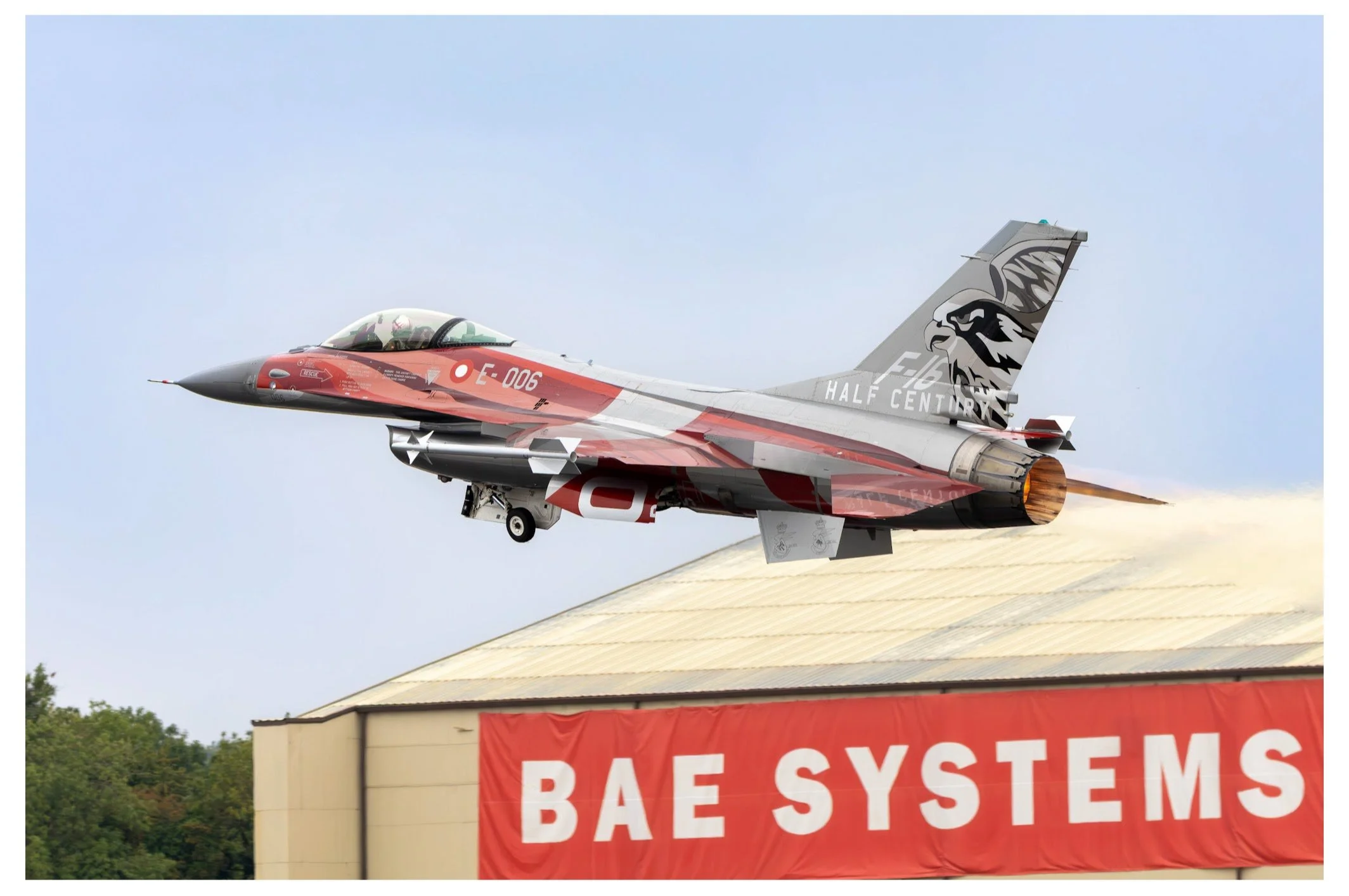 A fighter jet flying over a building with a red BAE Systems banner, with the jet painted with a tiger design on its tail and various military markings.