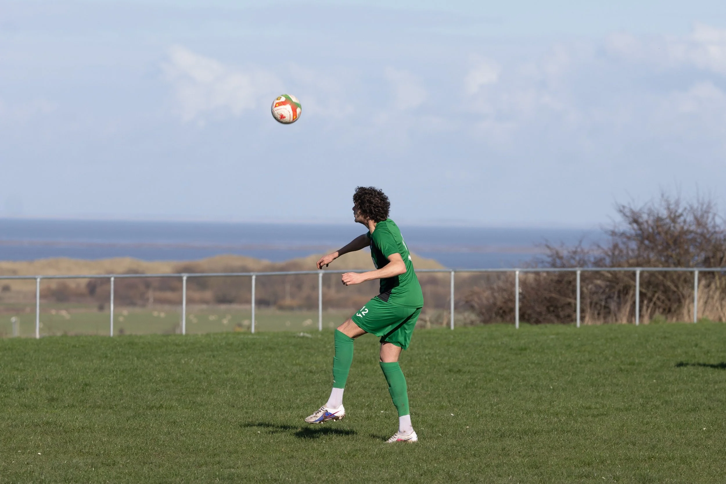 A soccer player in a green uniform is on a grassy field, preparing to kick a colorful soccer ball in mid-air.