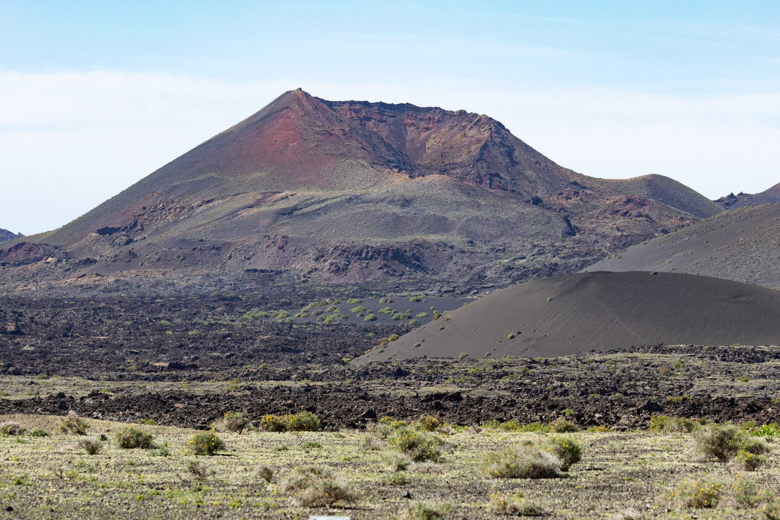 A volcanic landscape with dark, rocky terrain and a prominent volcanic peak in the background, under a light blue sky.
