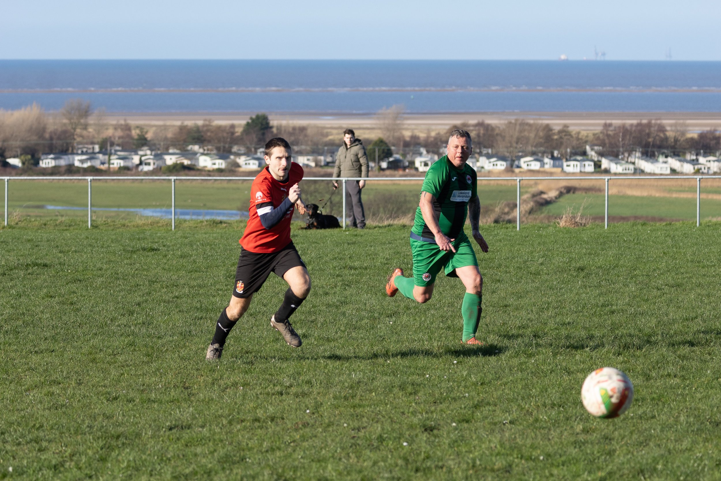 Two soccer players running on a grassy field, with a ball in front of them. One player is wearing a red jersey and black shorts, the other is wearing a green jersey and green shorts. A person with a dog is in the background.