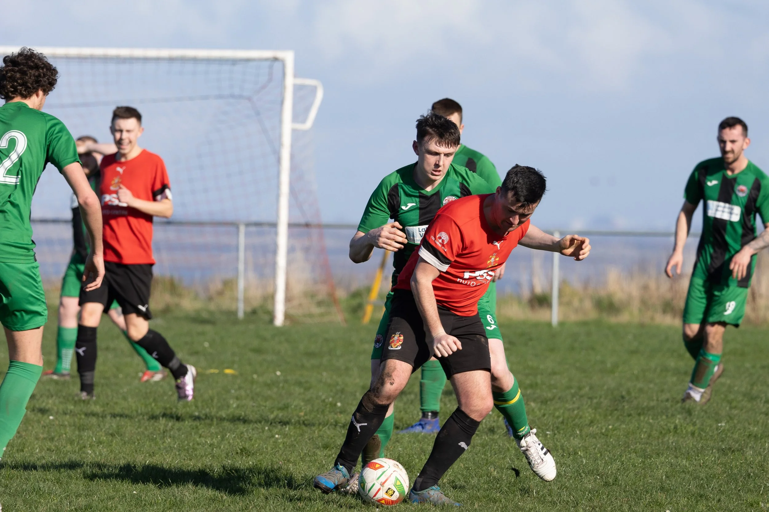 Soccer players in action on a grassy field during daytime, with two players from opposing teams competing for the ball near the goal, while other players observe or prepare to react.