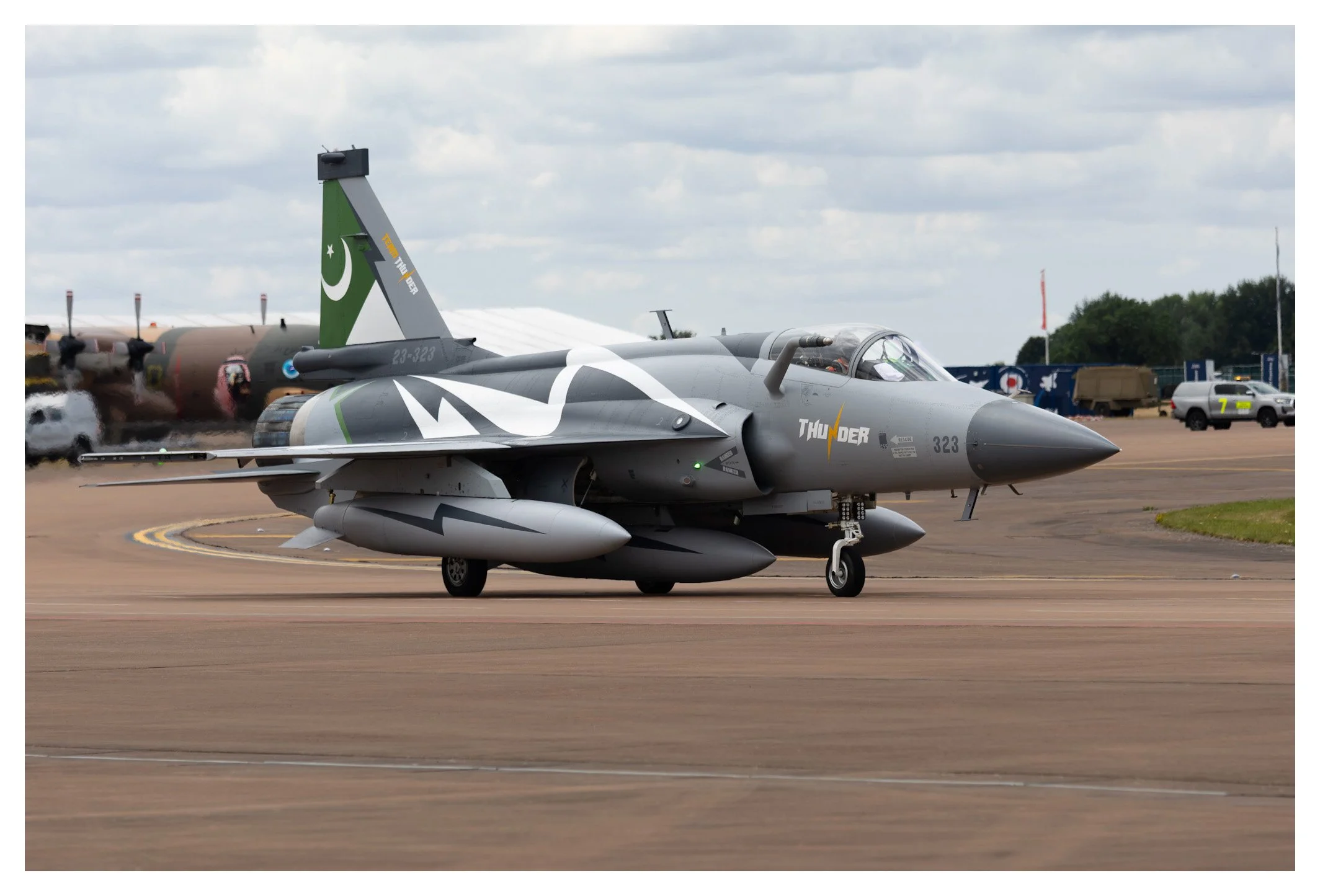 A military fighter jet on the runway, painted gray with a lightning bolt and the word 'THUNDER' on its side, against a cloudy sky background.