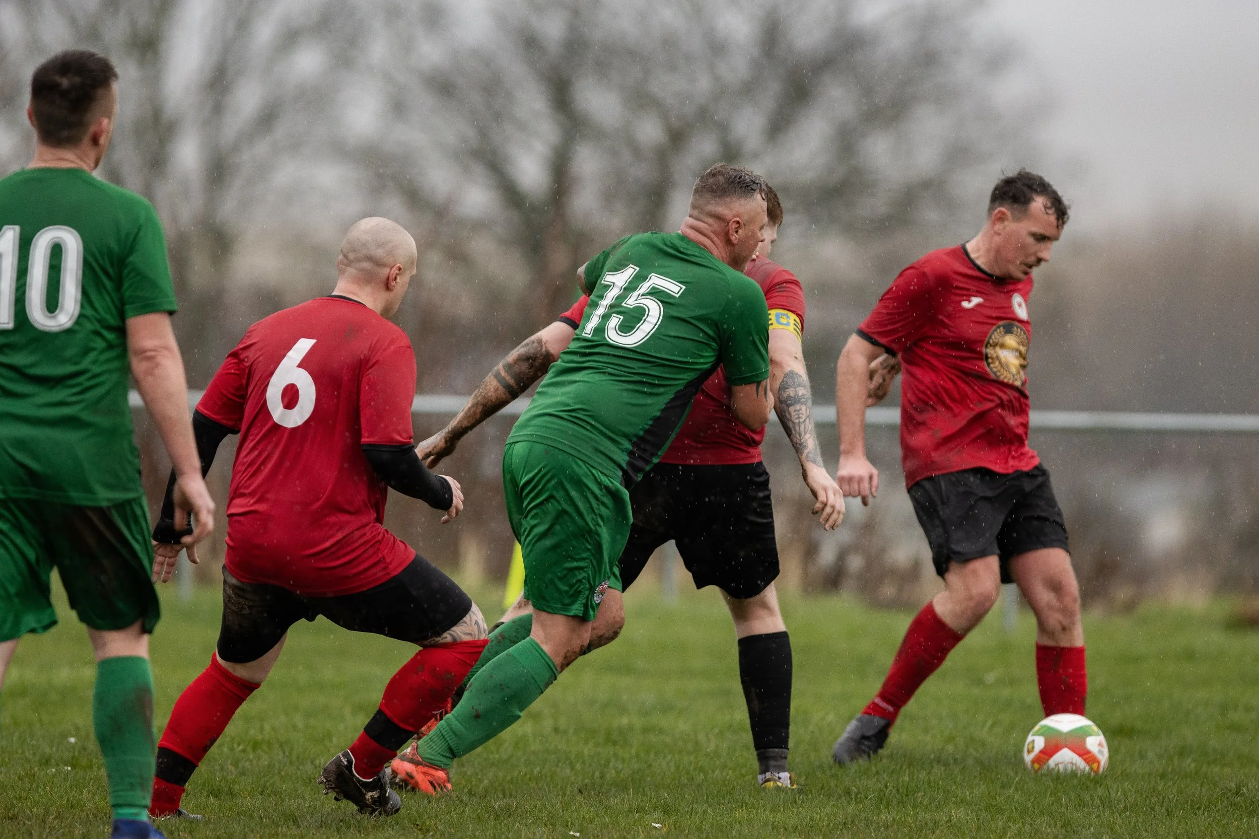 Soccer match with players in green and red jerseys, some wearing captain armbands, competing for the ball on a rainy day.