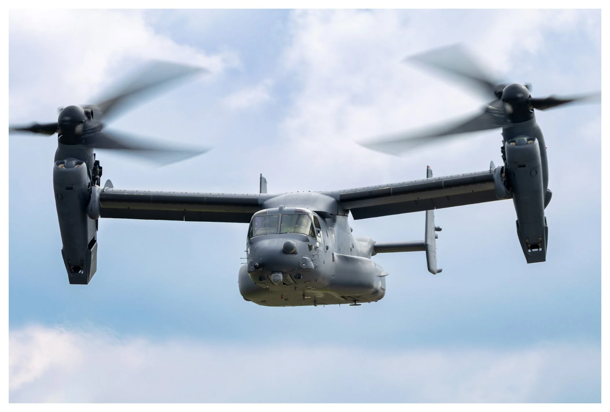 A gray V-22 Osprey military aircraft flying against a cloudy sky.