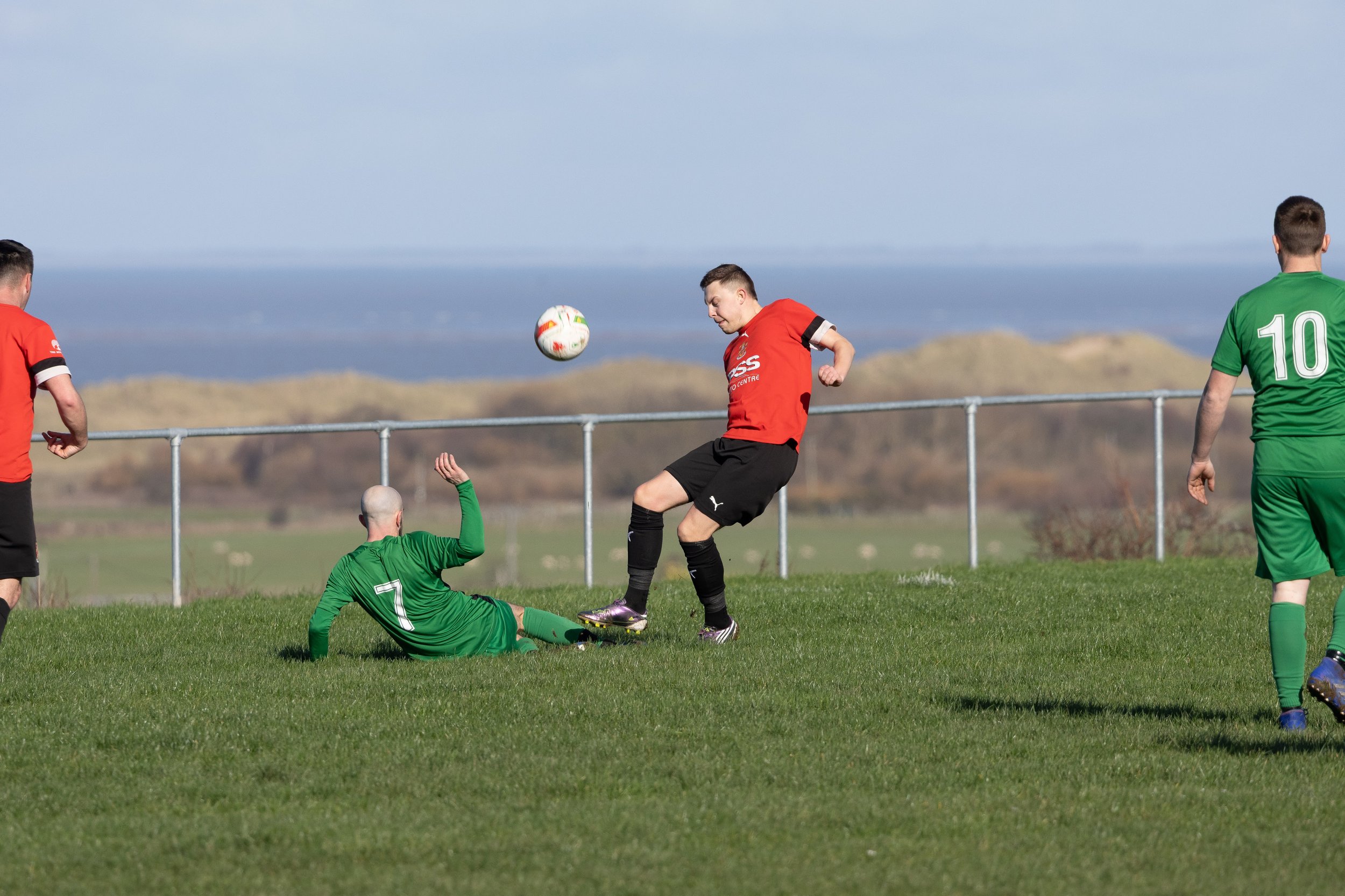 A soccer game with players in red and green uniforms on a grass field, with a goal and scenic landscape in the background.
