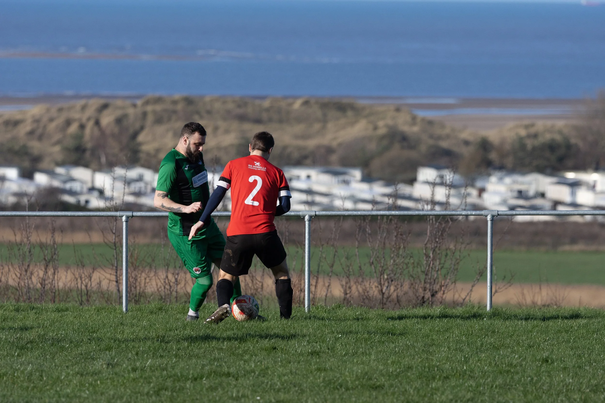 Two men playing soccer outdoors on a grassy field with a railing, hills, and bodies of water in the background.