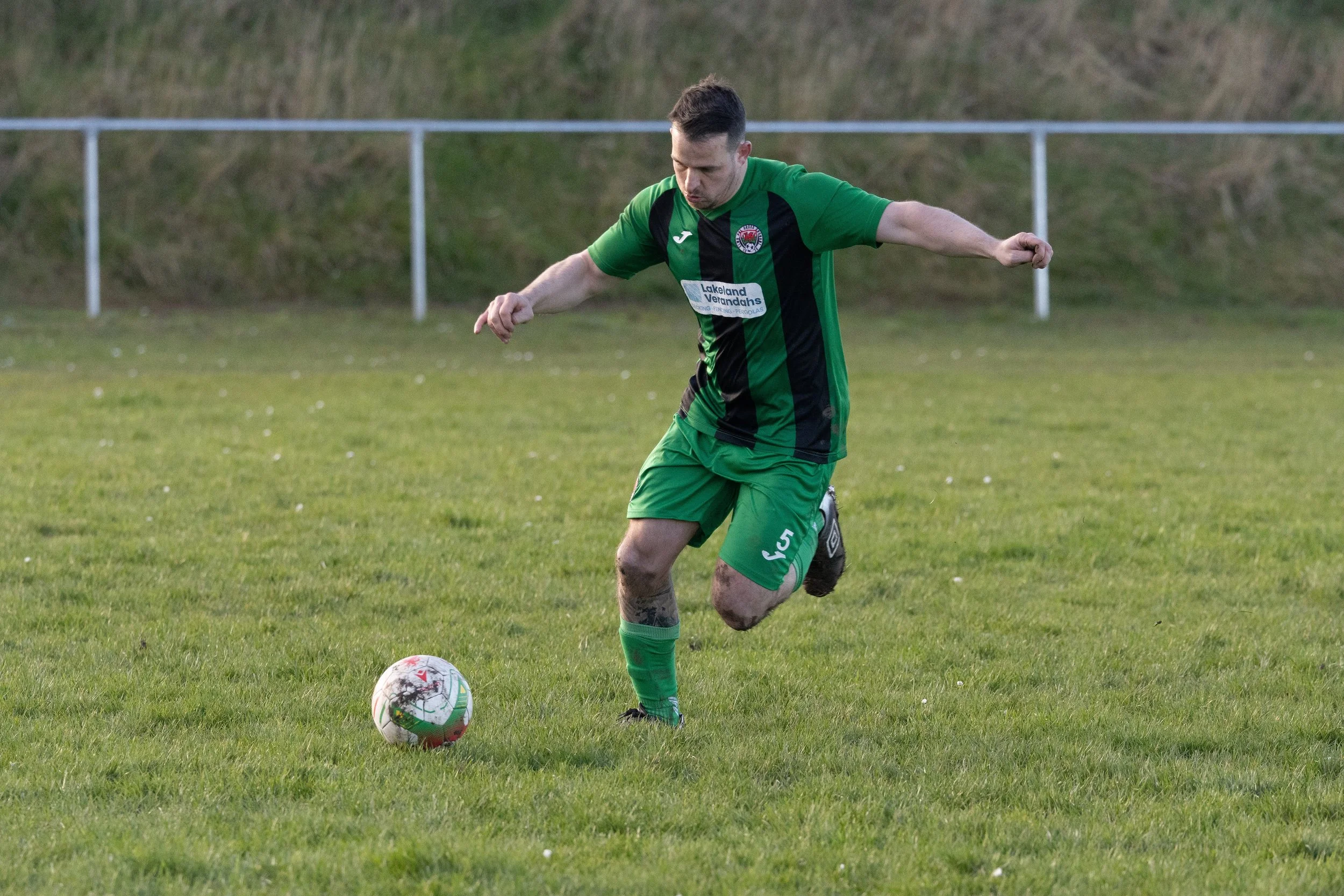 A male soccer player in a green and black uniform kicking a soccer ball on a grassy field.