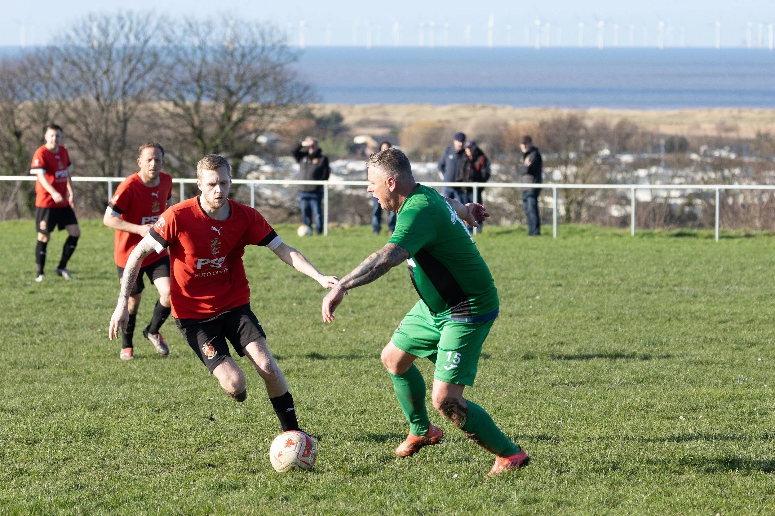 Soccer players in red and green jerseys competing for the ball on a grassy field, with spectators in the background and wind turbines in the distance.