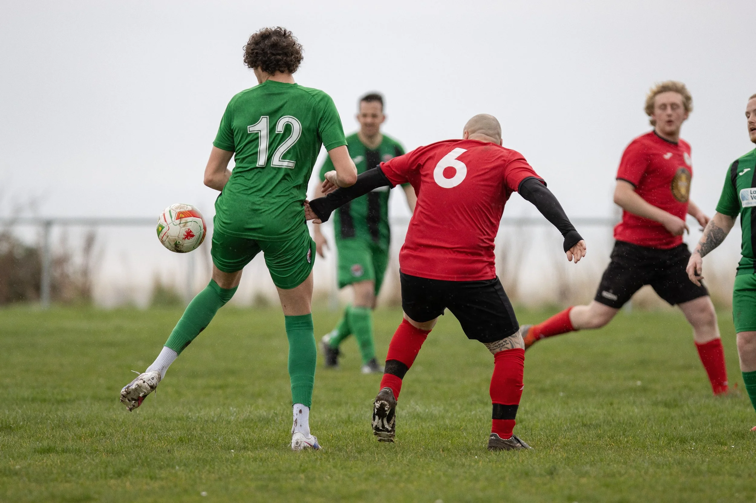 Soccer players competing for the ball during a match, with some players wearing green jerseys and others red jerseys, on a grassy field.
