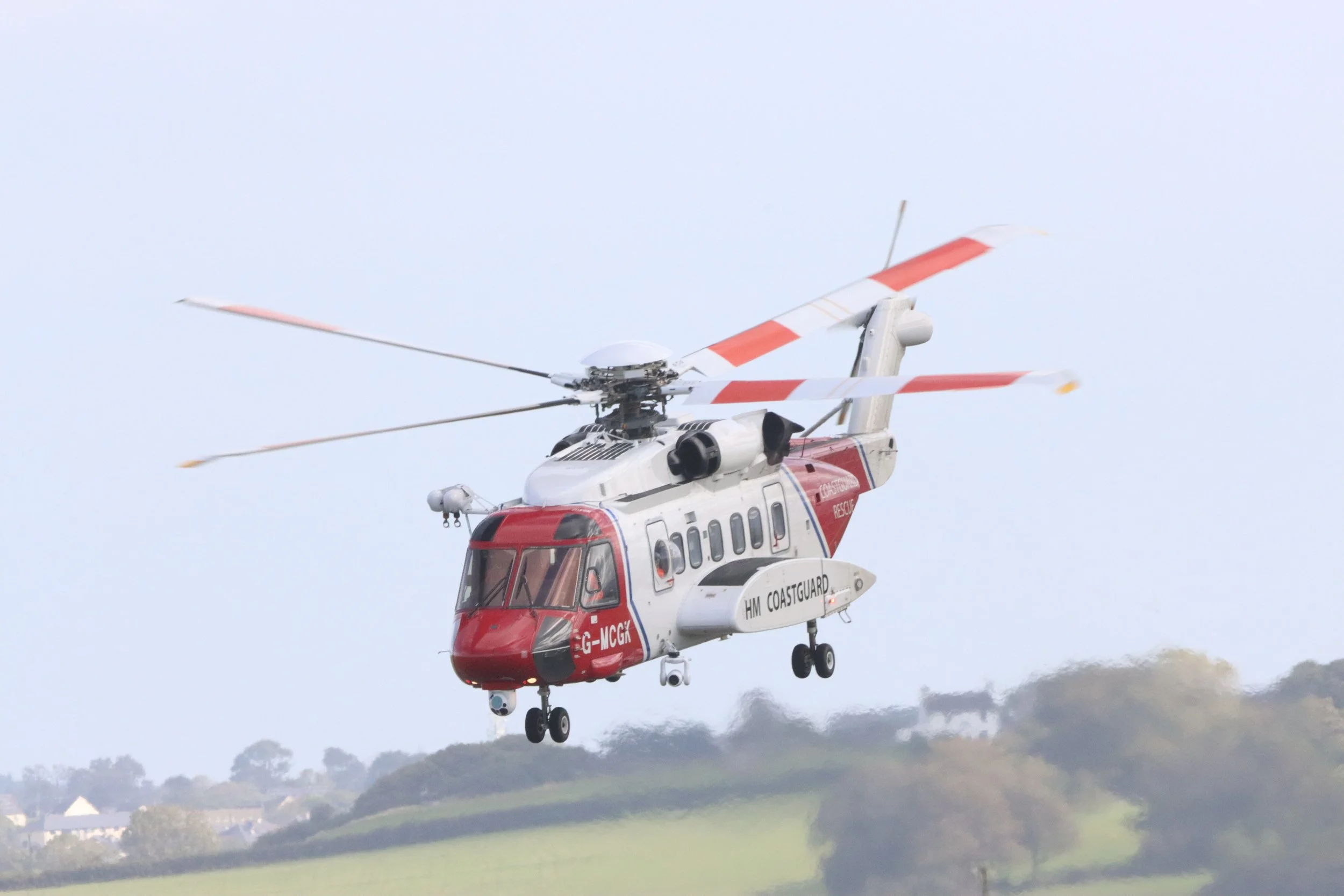 A rescue helicopter flying over a green landscape with trees and houses in the background.