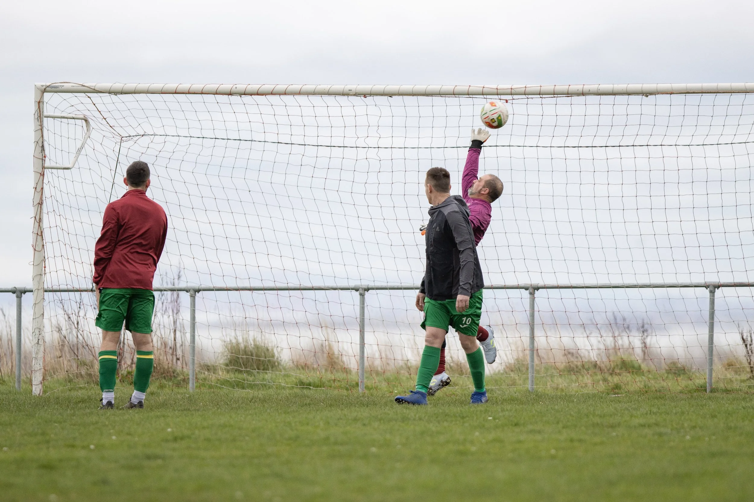 Soccer goal with three players, two wearing green shorts being goalkeeper, one in a maroon jacket and green shorts, on a grass field outdoors under cloudy sky.