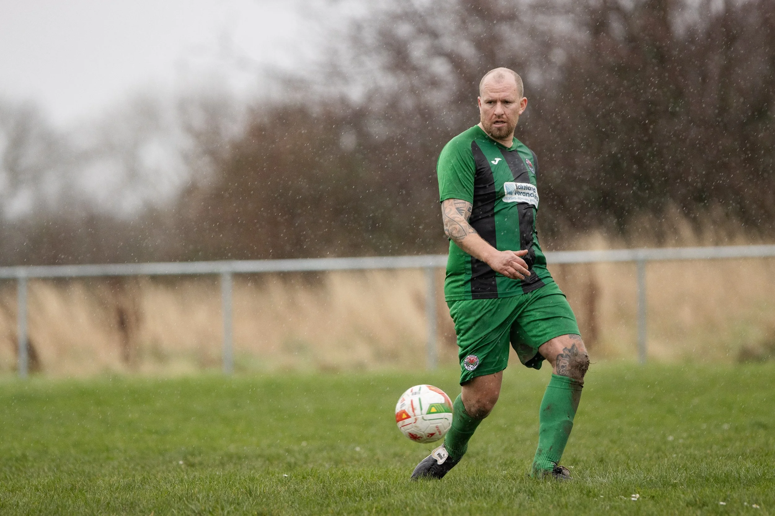 A man in a green and black soccer uniform playing soccer in the rain on a grassy field.
