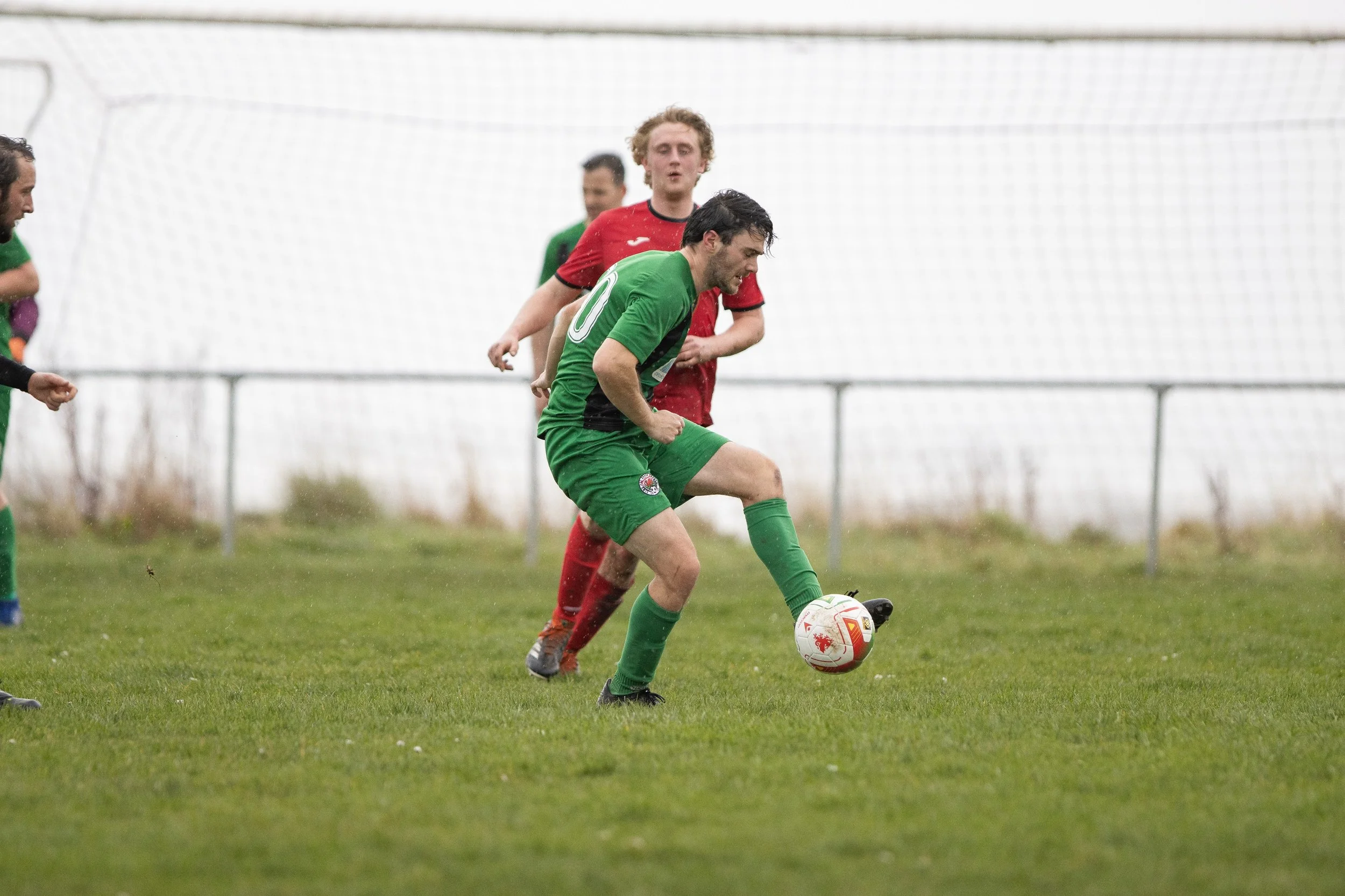 Soccer players in red and green jerseys competing for the ball on a grassy field, with a goal net in the background.