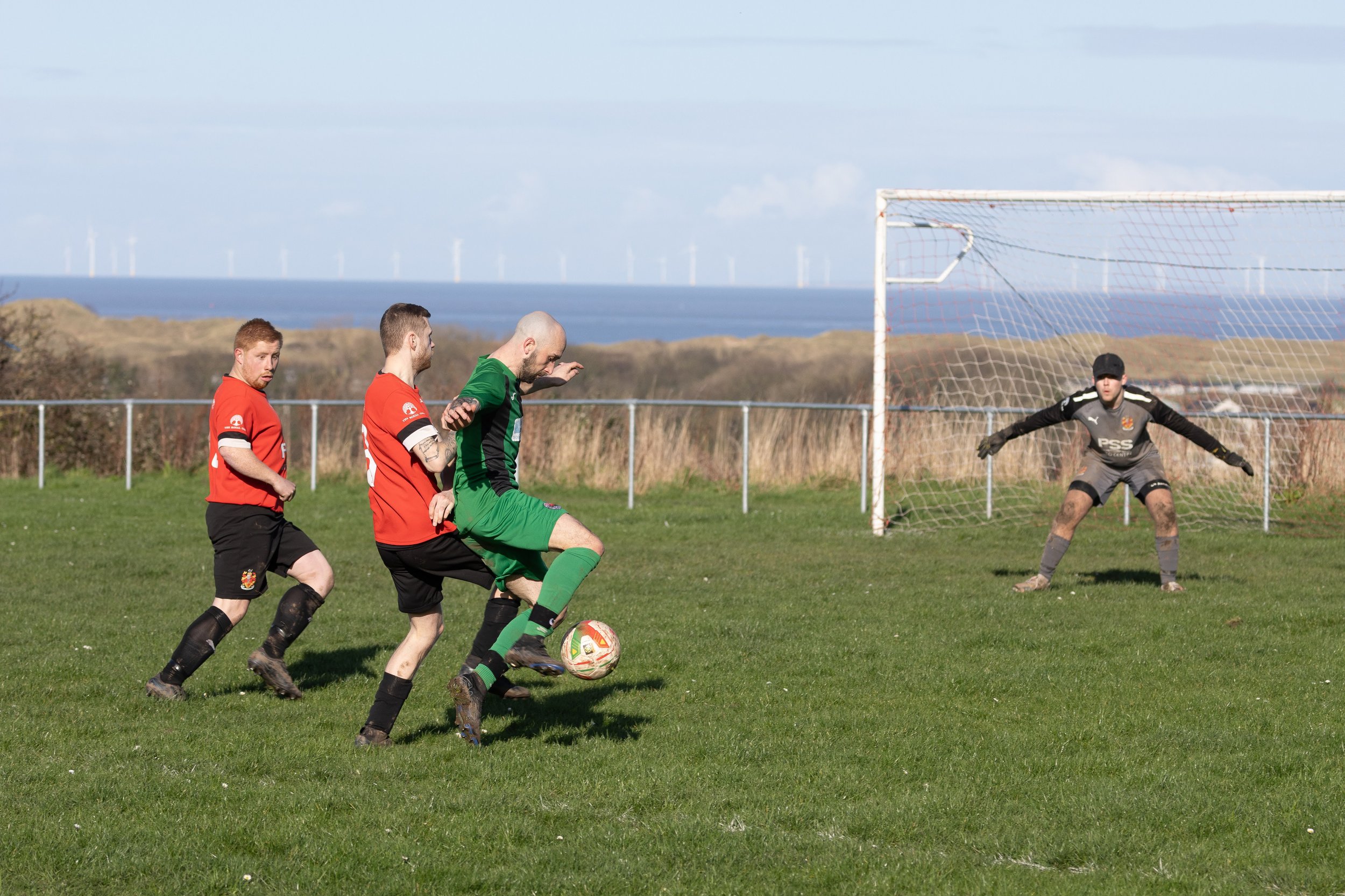 Soccer players competing for the ball near the goal, with the goalkeeper preparing to block the shot, on a field with a scenic background of the ocean and wind turbines.
