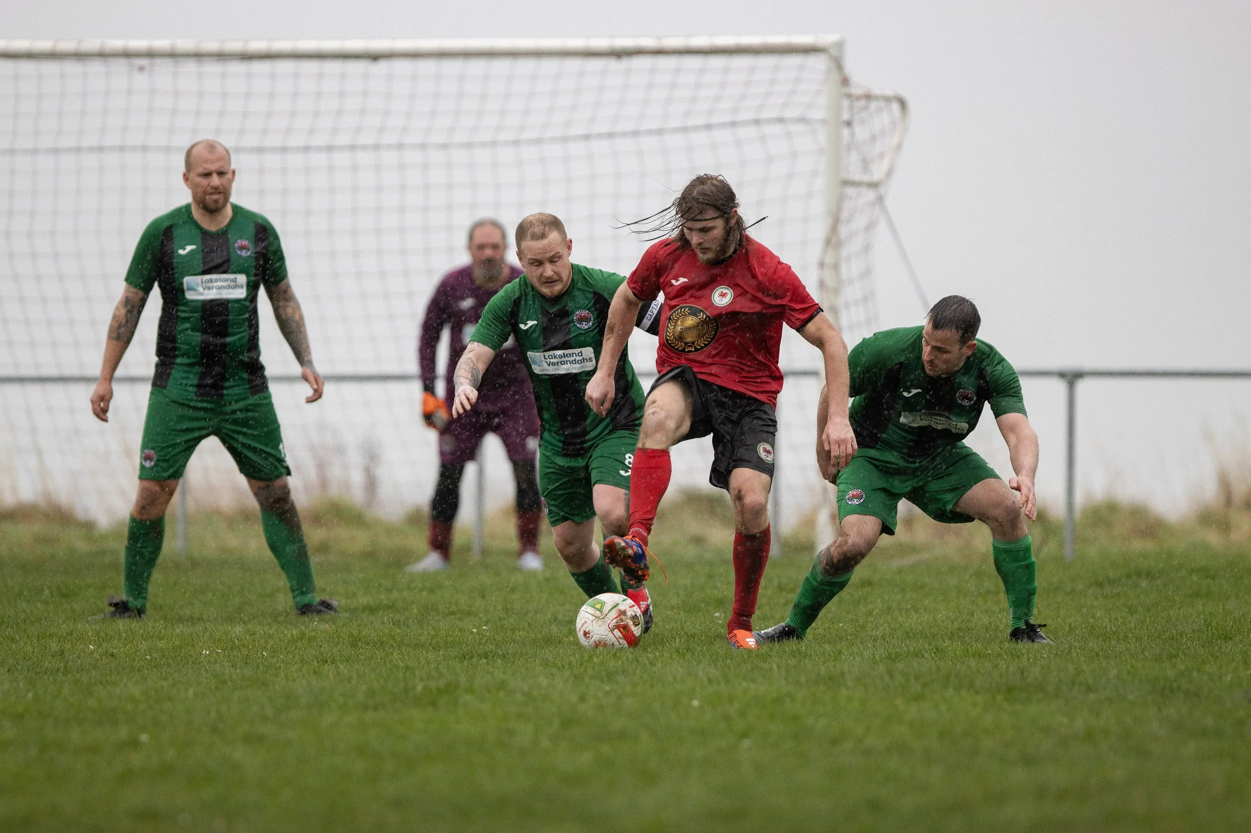 Four soccer players competing for the ball on a rainy day, with a goalpost in the background and wet grass.