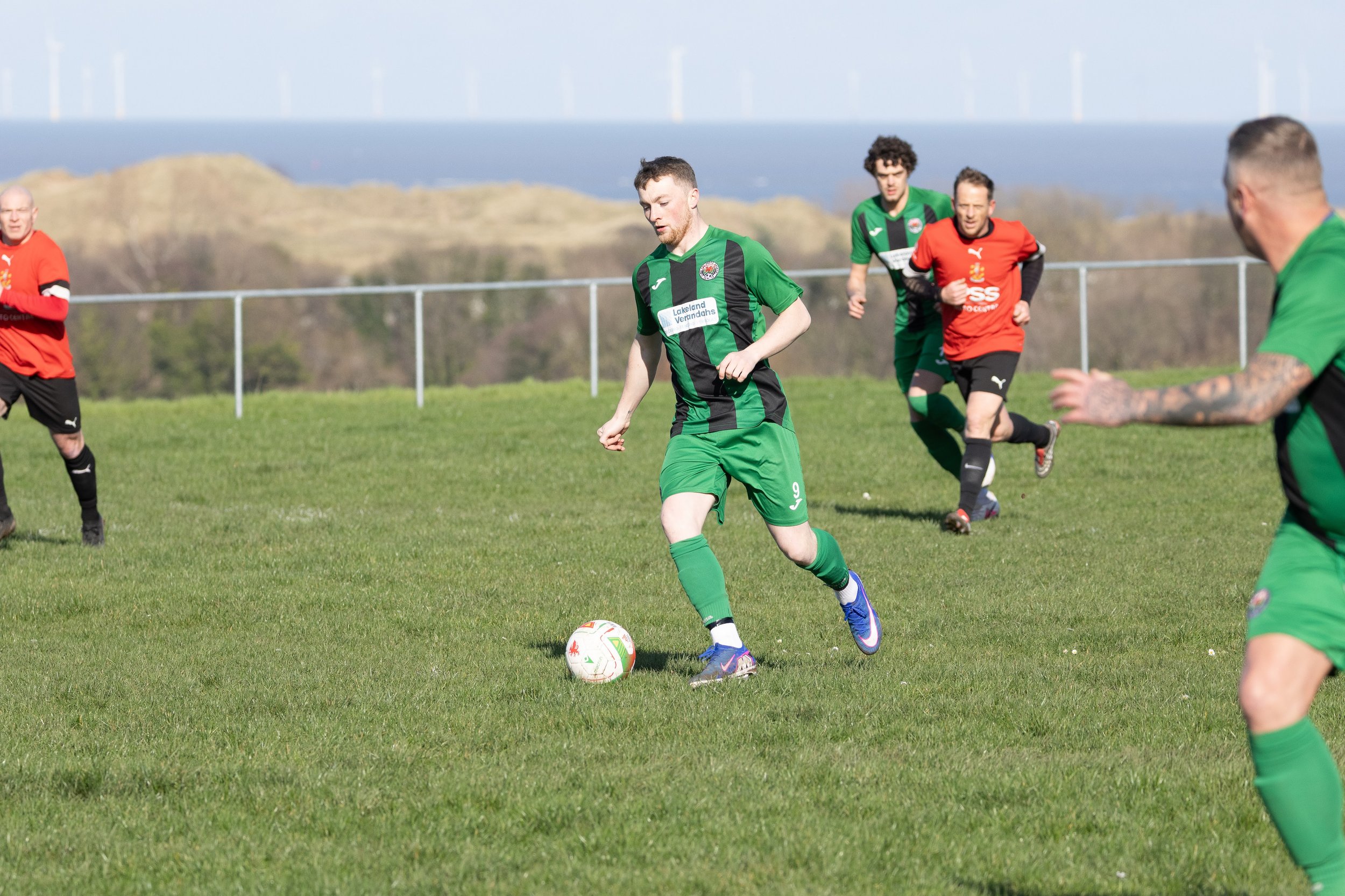 Soccer players in green and red uniforms playing on a grassy field with a fence and hills in the background.