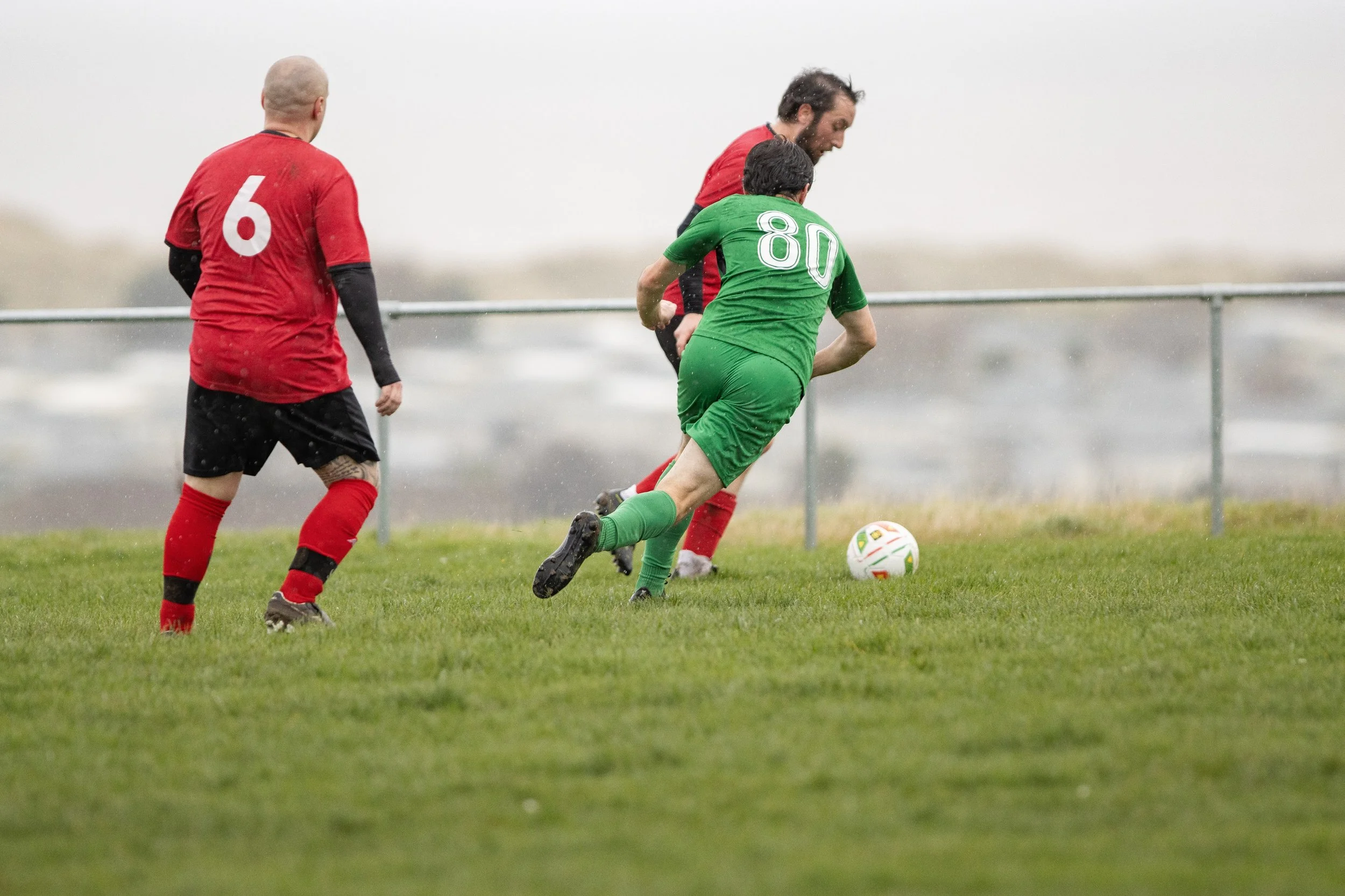 Soccer players in red and green uniforms competing for the ball on a grass field, rainy weather.