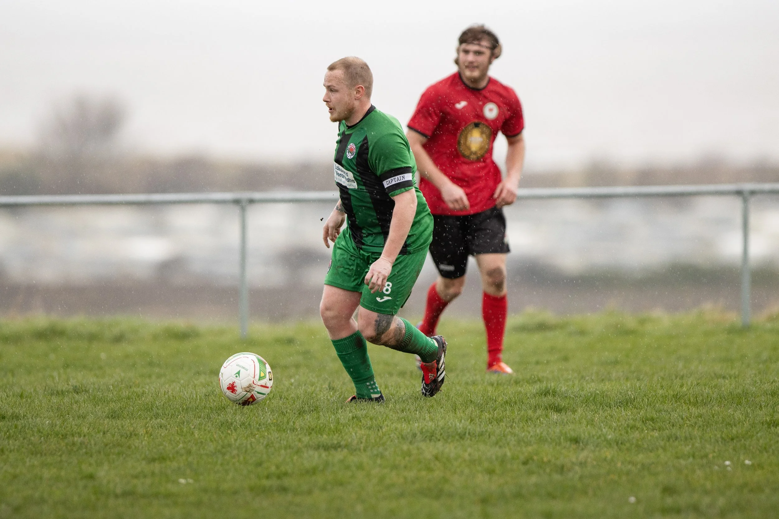 Two soccer players on the field during a match, with one in a green uniform controlling the ball and the other in a red uniform in the background, on a rainy day.