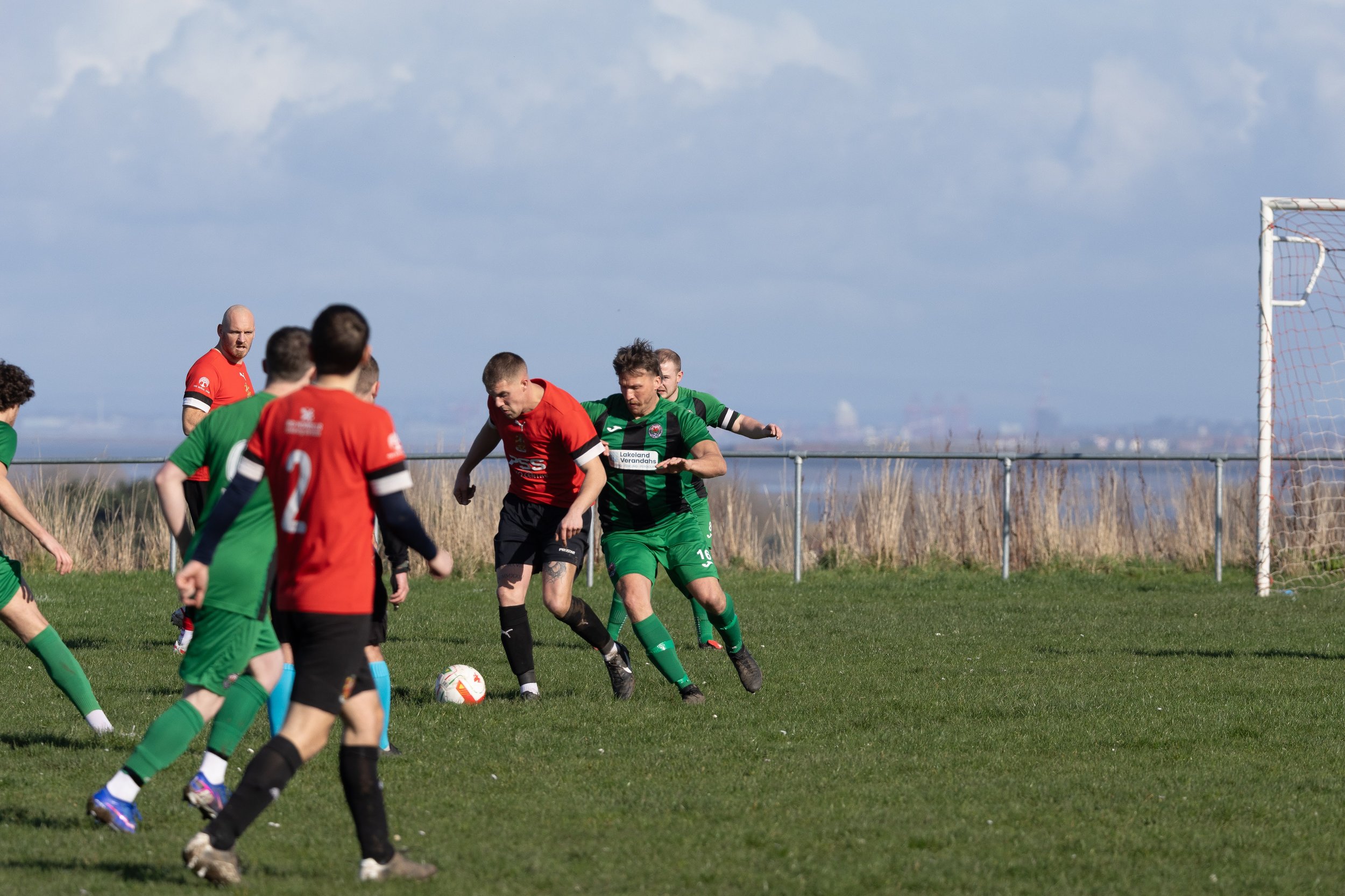 Soccer match with players fighting for the ball on a grassy field, some players in red and others in green jerseys, with a goalpost in the background and a cloudy sky overhead.