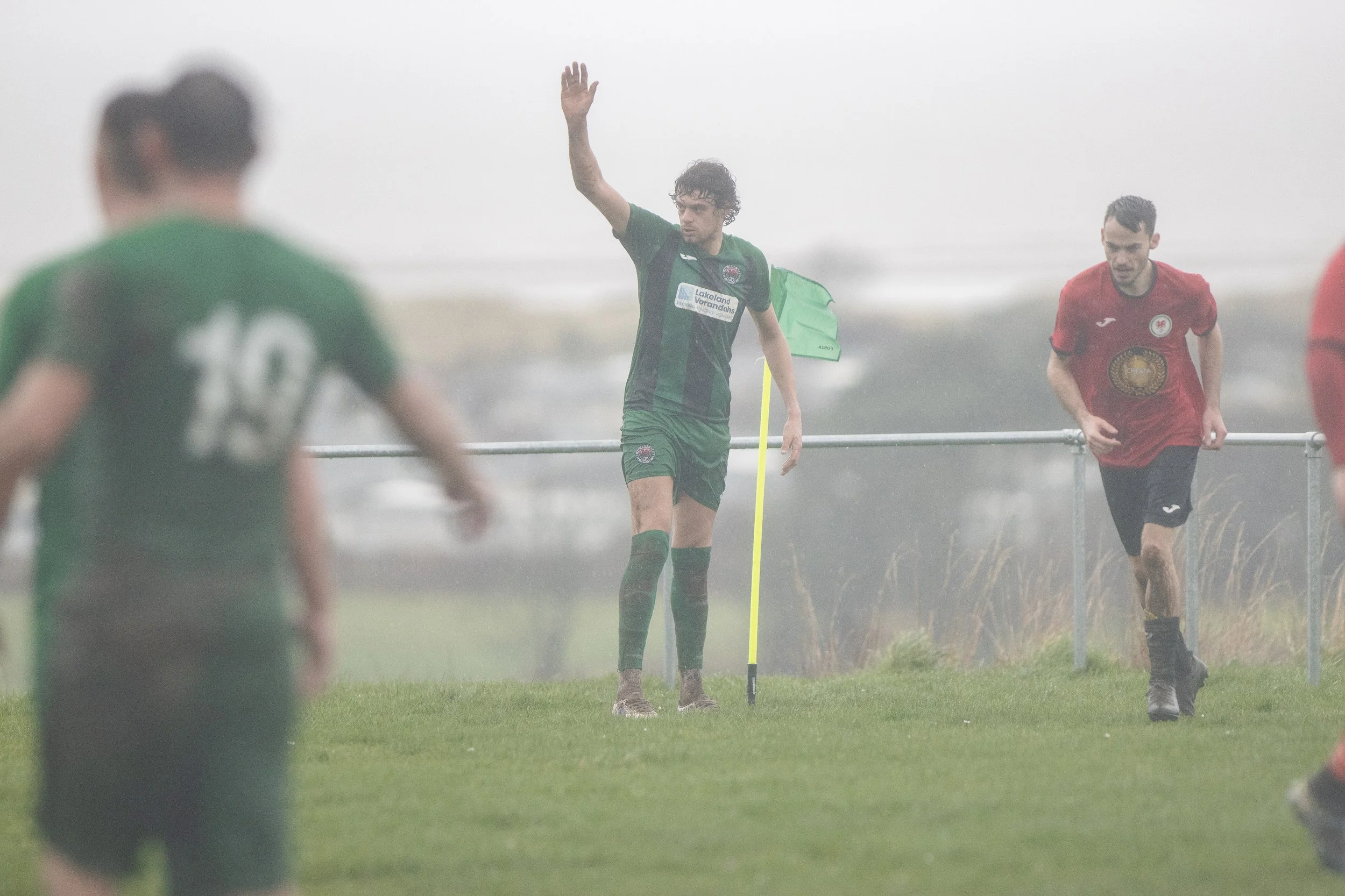 Soccer players on a foggy field, with one player holding a corner flag and others running. The players wear green and red uniforms.