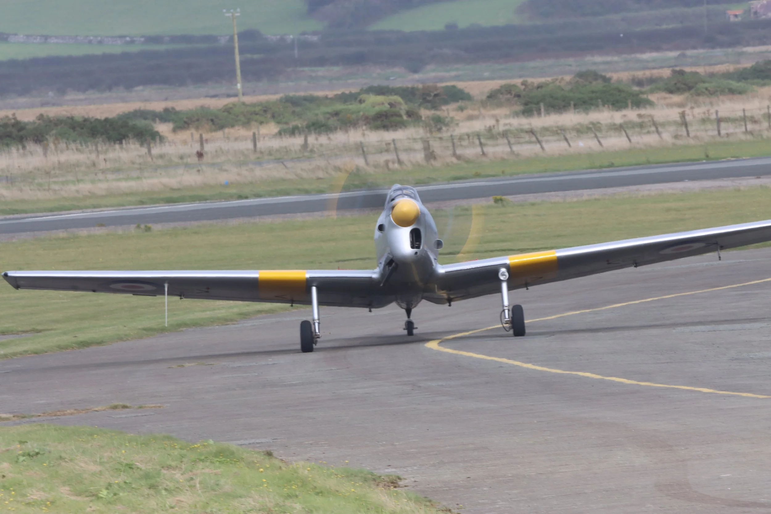 A silver vintage WWII aircraft with yellow markings on the wings, taxiing on a runway.
