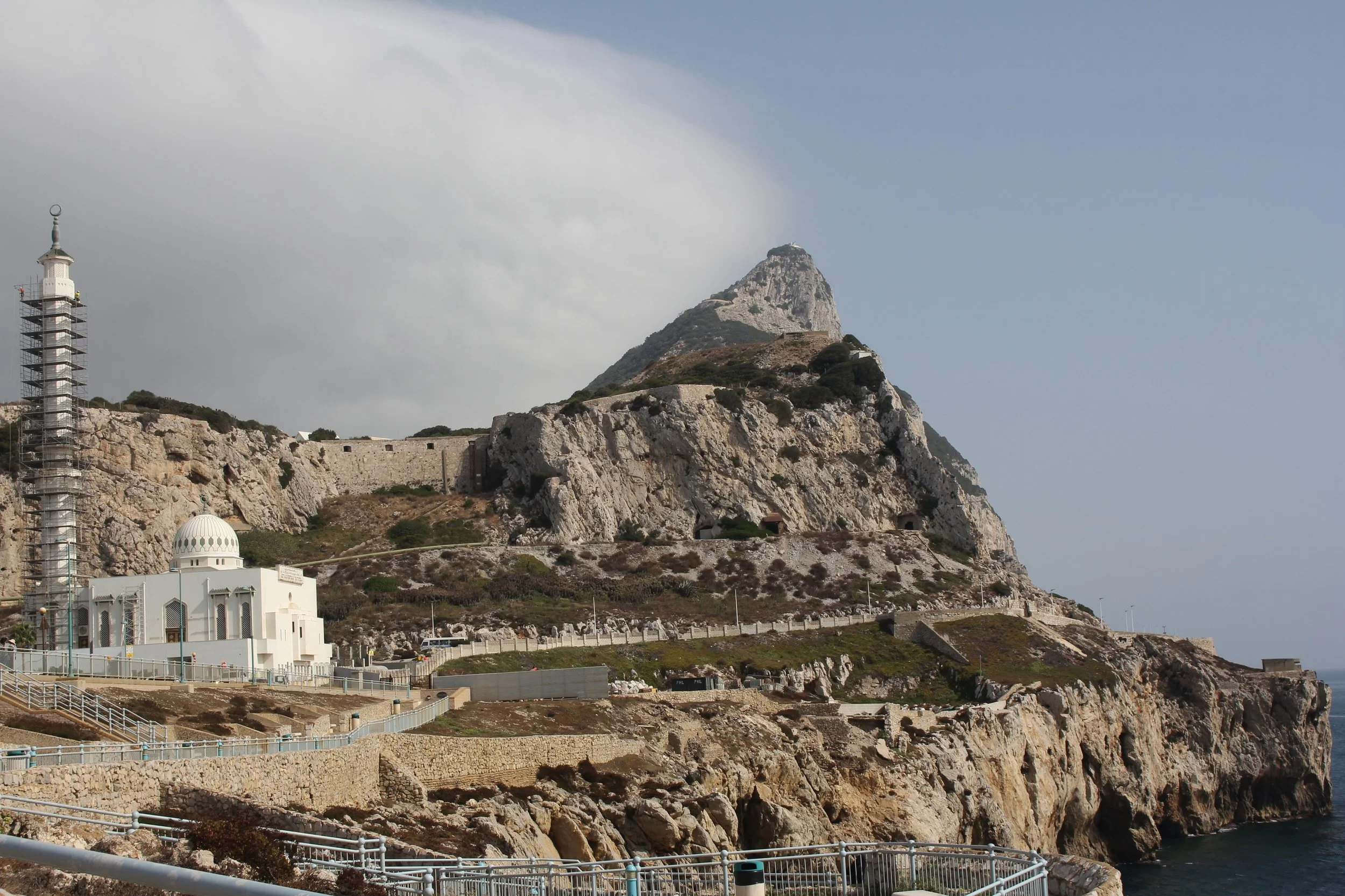 A rocky hillside with a lighthouse on the left, a white mosque-like building, and a fortress wall along the top of the hill. The sky is partly cloudy with blue visible.