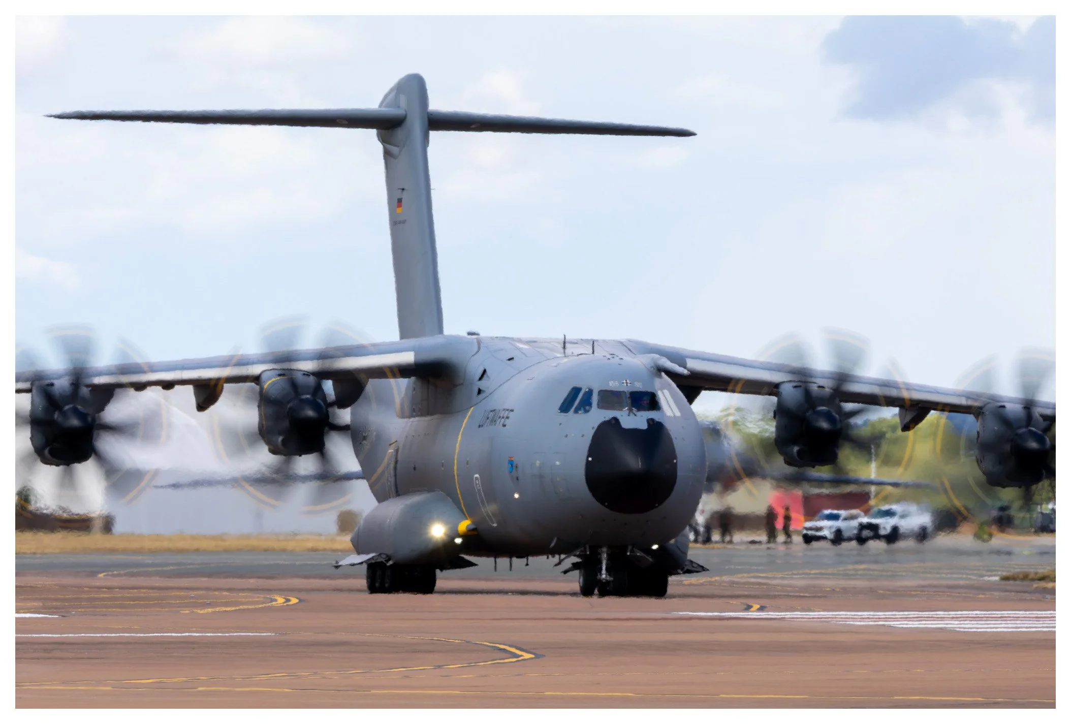 Military cargo aircraft on runway with spinning propellers, gray color, large size, and high wings, with several vehicles and people in the background.
