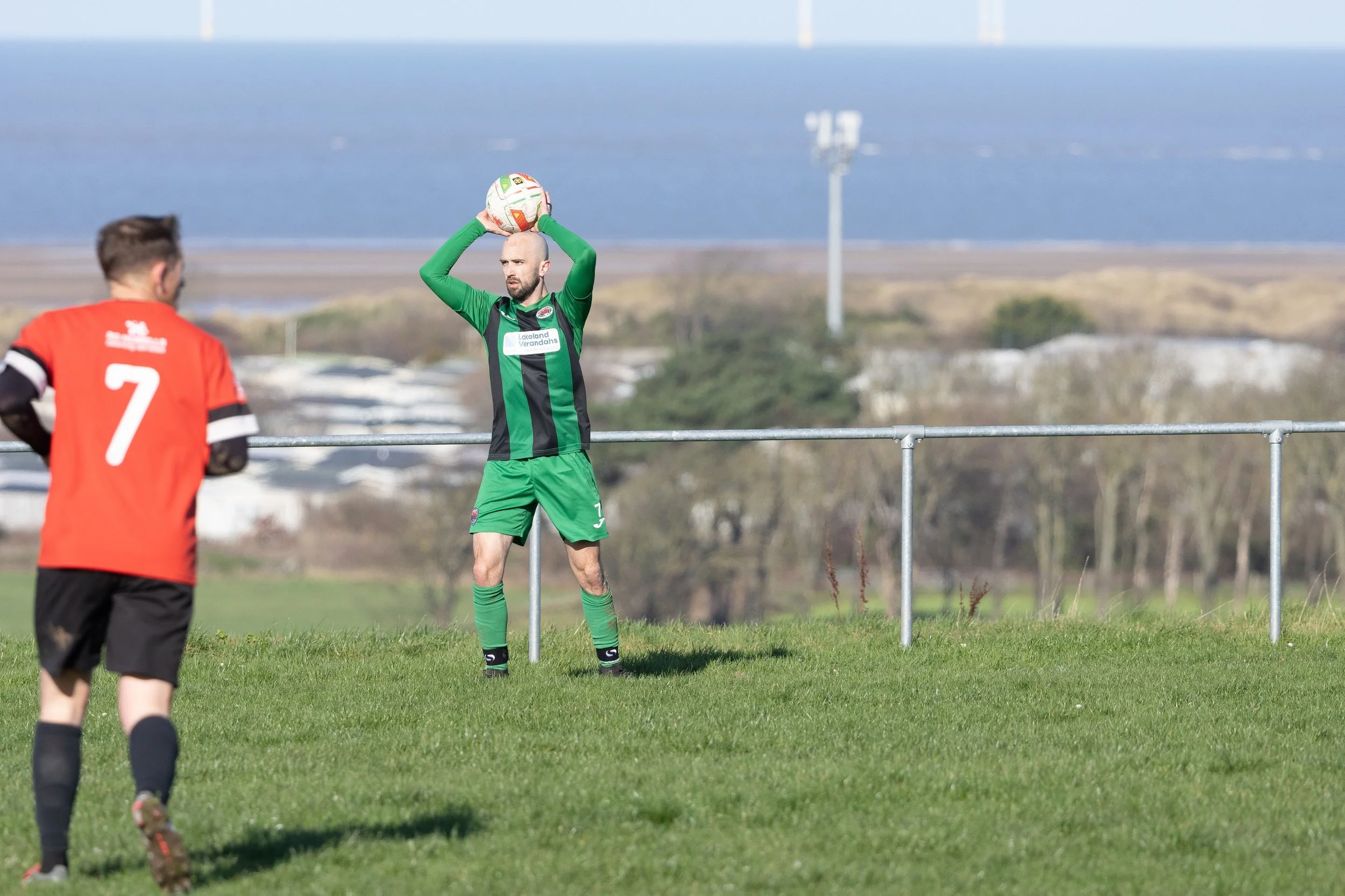 A soccer player in a green uniform holding a soccer ball above his head, preparing to throw-in during a game on a grassy field, with another player in a red and black uniform observing nearby and a scenic background of ocean and hills.