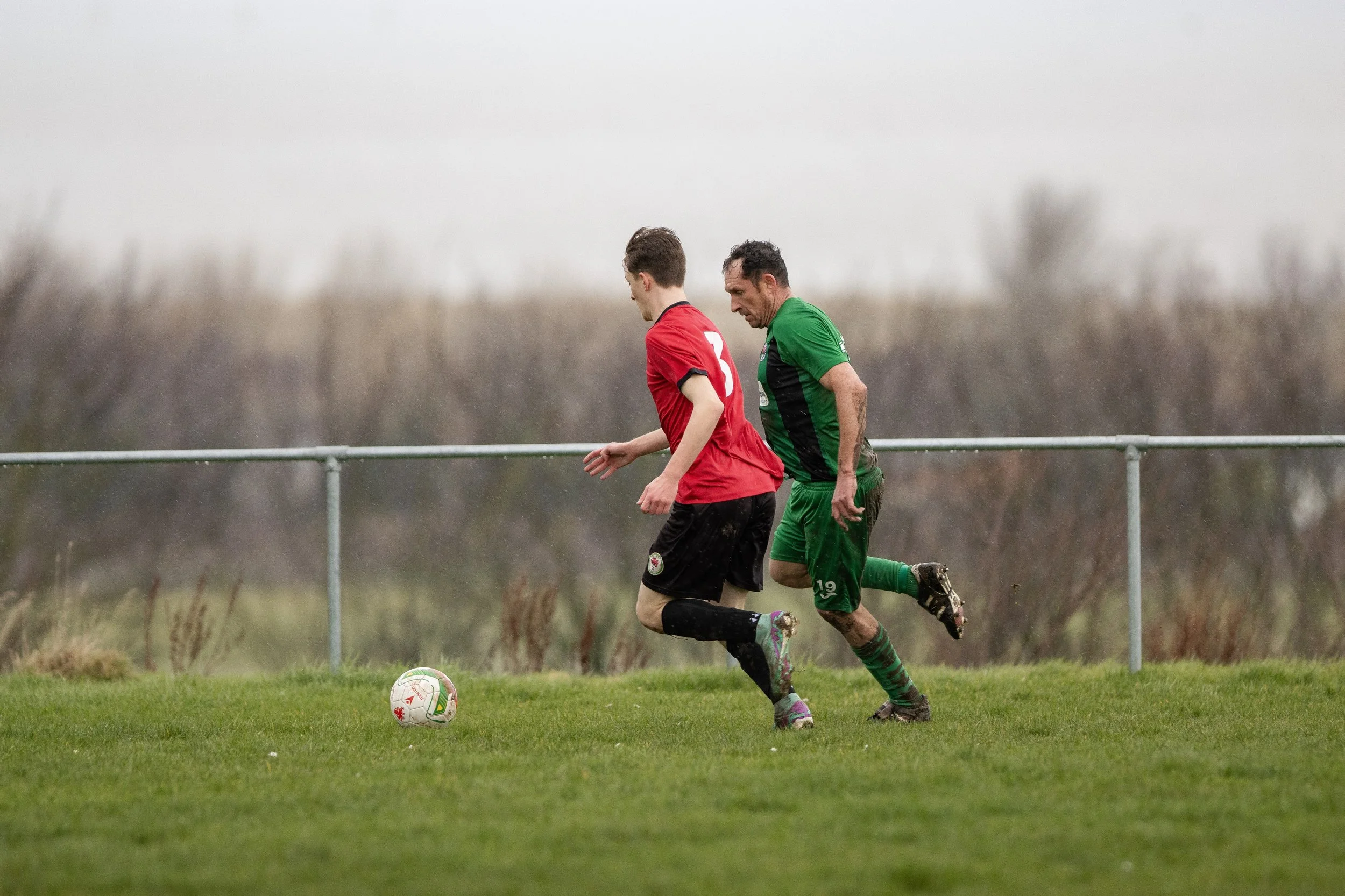 Two soccer players, one in a red jersey and the other in a green jersey, compete for the ball on a grassy field during a rainy day.